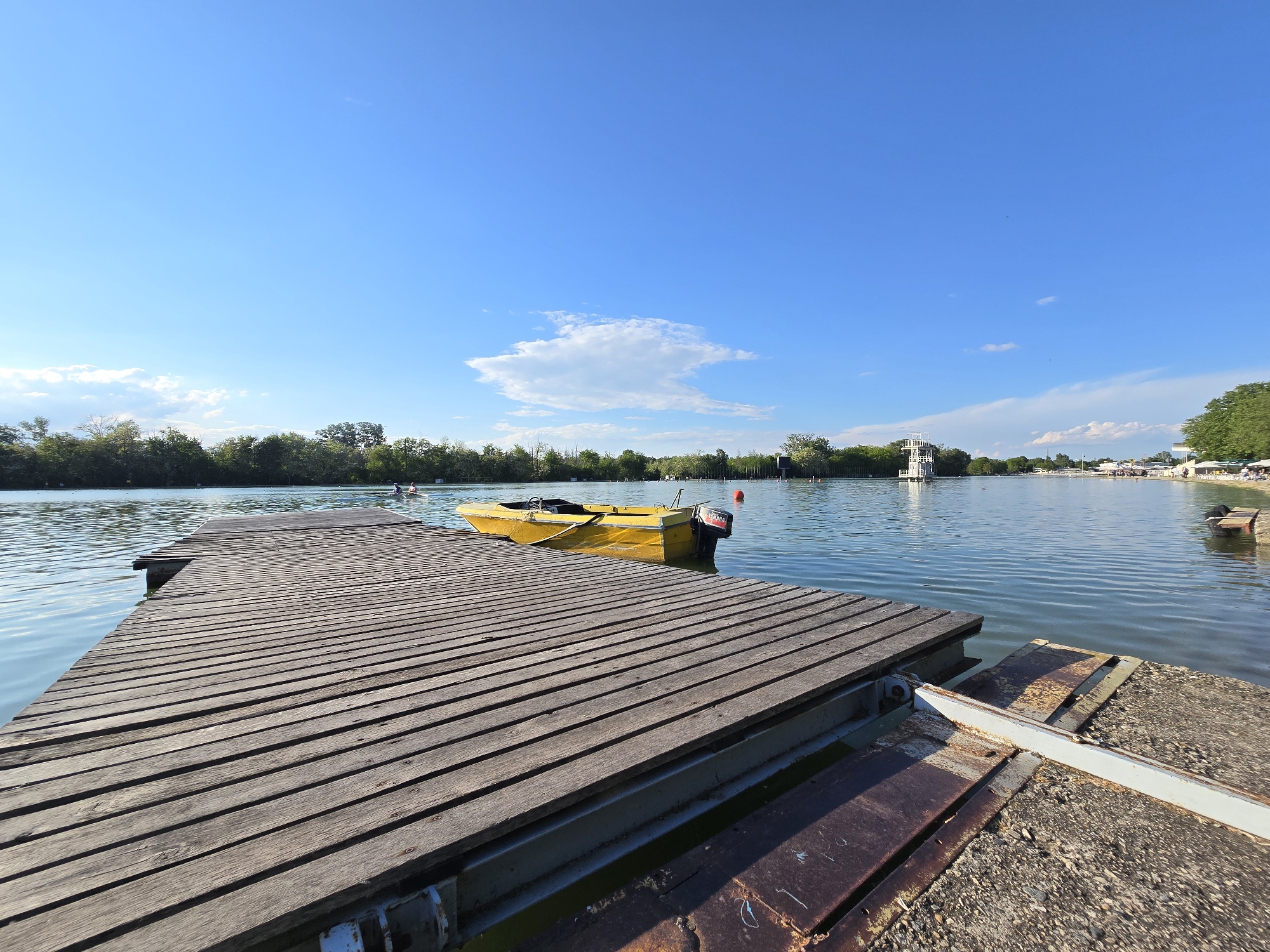 The photograph shows a yellow boat tied to a wooden pier on a lake in Unknown. The pier is made of dark wood and is connected to a concrete and rusted metal structure. The lake is surrounded by trees. In the distance, there is a diving platform and a beach with people. The sky is blue with a few clouds.