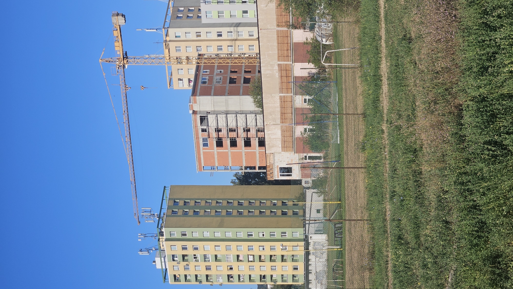 The image depicts a construction site in the city of Prešov, Slovakia. A large yellow tower crane is prominently featured, extending horizontally across the sky. In the background, several multi-story buildings are visible. One tall residential building on the left is painted in yellow and olive green, with numerous windows. To its right, a partially constructed building with exposed brickwork and scaffolding indicates ongoing construction. Further to the right, another residential building is partially visible. In the foreground, a sports field, possibly a soccer field, is enclosed by a wire fence. The field appears to be a mix of grass and dirt. The sky is a clear, bright blue. The overall scene suggests urban development and ongoing construction activity in the city.