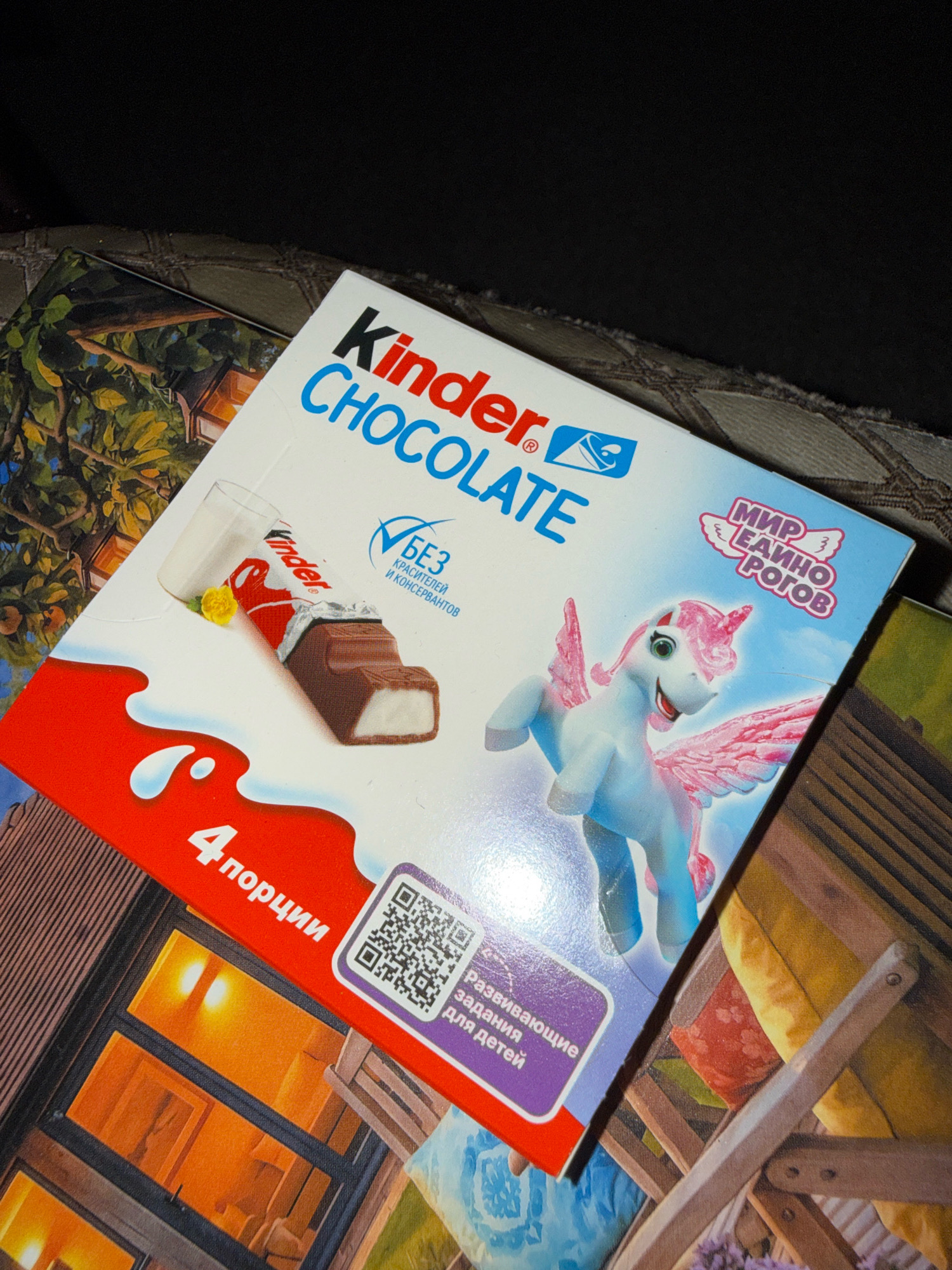 A white Kinder Chocolate box with a winged unicorn and Russian text is placed on a textured surface, possibly a rug or blanket. The box is angled, showing the front and a portion of the top. The text on the box includes "Kinder CHOCOLATE", "БЕЗ КРАСИТЕЛЕЙ И КОНСЕРВАНТОВ" (without dyes and preservatives), "МИР ЕДИНОРОГОВ" (world of unicorns), and "4 порции" (4 portions). A QR code and text "Развивающие задания для детей" (educational tasks for children) are also visible on the bottom right of the box. The background features a blurred image of what appears to be a wooden structure with windows and a portion of a patterned fabric. The lighting suggests indoor conditions. There are no people visible in the image.