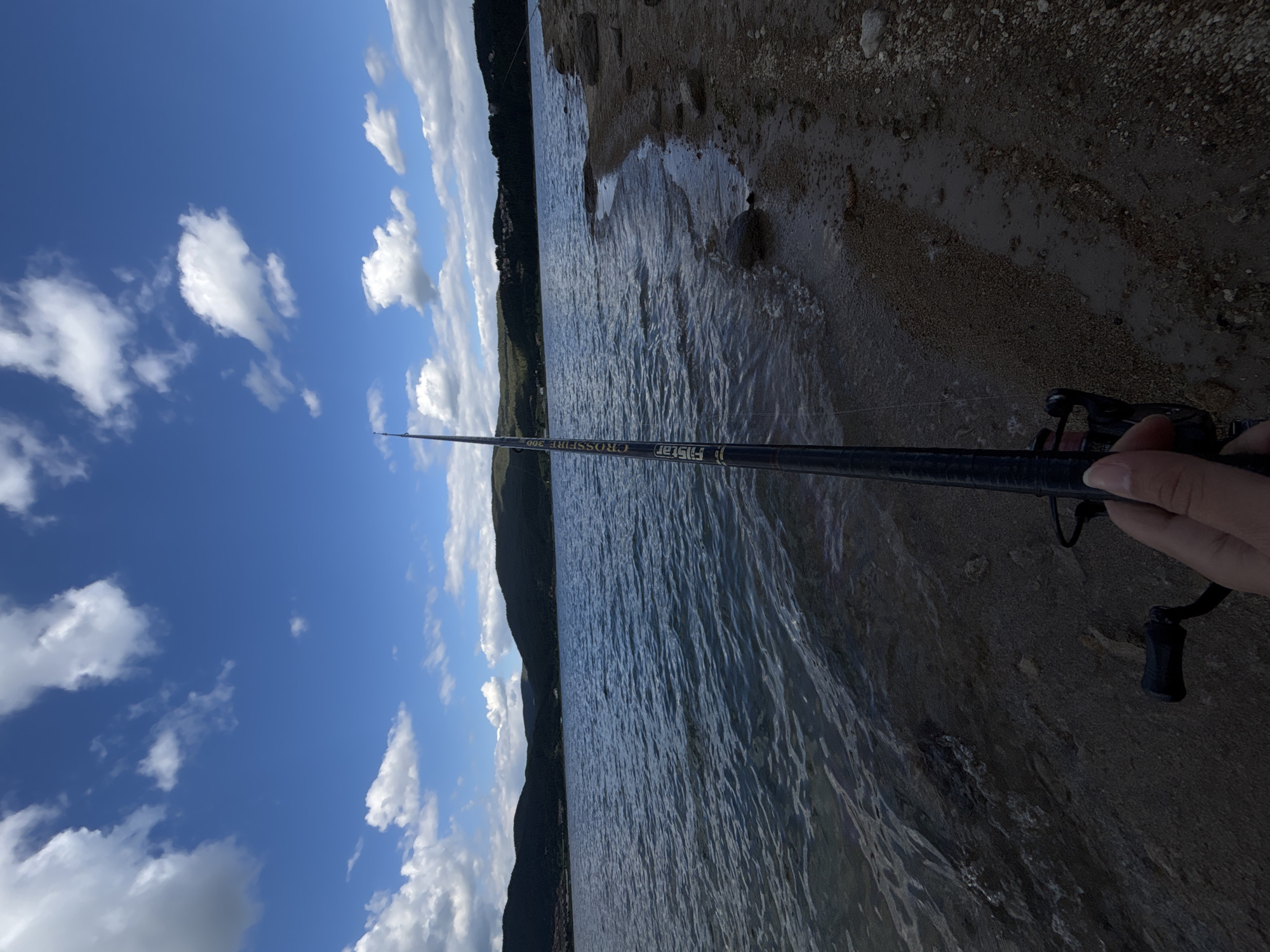 The image depicts a person's hand holding a fishing rod by the water's edge, presumably on a beach or shoreline composed of sand and pebbles. The fishing rod is black and has visible text on it that reads "FISHSTER CROSSFIRE 300". The water appears calm with gentle ripples, reflecting the bright blue sky filled with scattered white clouds. In the background, a dark, tree-covered hillside slopes down towards the water, with glimpses of what might be buildings or structures along its base. The overall scene suggests a peaceful, outdoor activity, likely fishing, on a clear, sunny day. The orientation of the image is vertical, with the sky occupying the left portion and the water and shore the right.