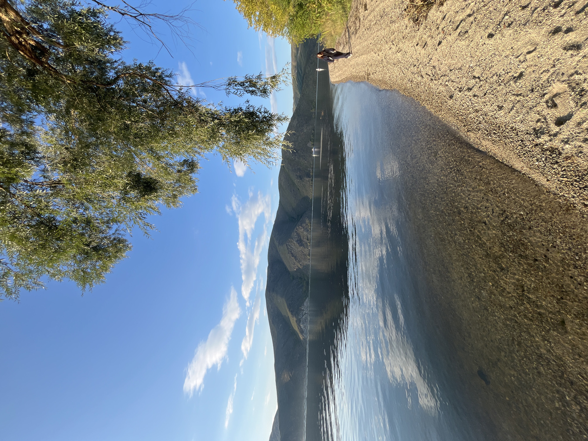 The image depicts a serene natural landscape by a body of water, likely a river or lake, in or near the city of Oskemen, Kazakhstan. The scene is dominated by a wide expanse of calm water reflecting the blue sky and scattered white clouds, with a range of verdant, tree-covered hills forming the distant horizon. The foreground features a narrow, sandy and pebbly shoreline. A lone woman, dressed in dark clothing, walks along the shoreline, her back to the viewer. Overhanging branches of a leafy tree frame the top portion of the image, casting dappled sunlight on the scene. In the water, two small white boats are visible in the distance. The overall impression is one of tranquility and natural beauty, possibly during a late afternoon or early morning given the lighting.