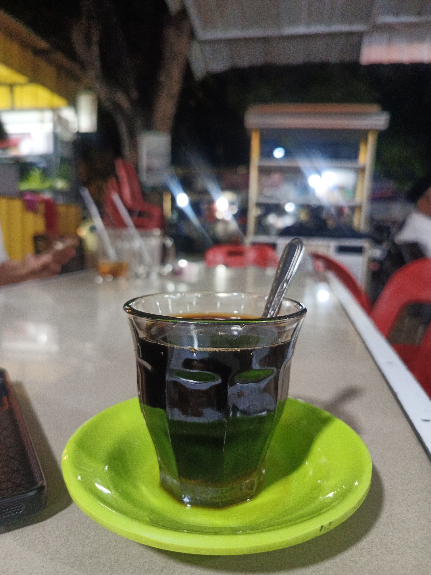 The image presents a cup of dark liquid, possibly coffee or tea, served in a clear glass on a bright green saucer. A spoon is inside the glass. The cup and saucer sit on a table, with a black phone visible to the left. In the blurred background, there is a partially visible person using a phone, some additional clear drinking glasses, red plastic chairs, and food stalls illuminated with bright lights, indicating an outdoor or street-side cafe setting. The image was likely taken at night. Considering the prompt, the location is likely in Lhokseumawe, Indonesia.
