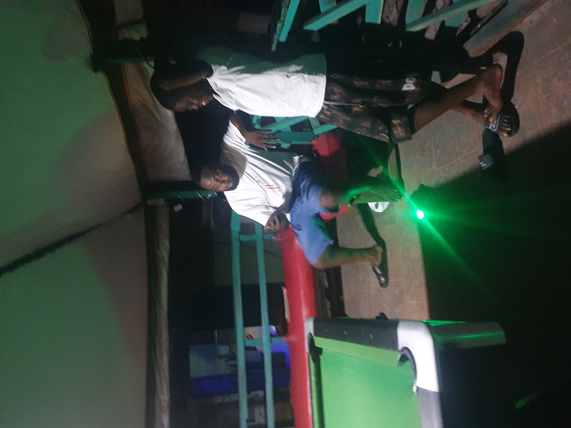 The image depicts a group of young men in what appears to be a dimly lit outdoor setting, possibly a bar or a recreational area, as suggested by the presence of a pool table in the foreground. The scene is illuminated by a bright green light, casting distinct shadows and creating a vibrant, almost surreal atmosphere.
Three men are clearly visible. One man, wearing a white t-shirt, is seated and looking down. Another man, also in a white t-shirt and dark shorts, is standing and appears to be interacting with the seated man. A third man, wearing a blue shirt and dark shorts, is partially visible, leaning on a red pole. His legs and feet are also visible. All three men appear to be young adults.
The pool table is green with black and white trim. The surrounding area is dark, with hints of green walls or structures. The overall impression is that of a relaxed evening gathering. The time of day is likely night, given the darkness and the artificial lighting. There are no clear indications of weather conditions.
The men's expressions are difficult to discern clearly due to the lighting and image quality, but the interaction seems casual and friendly. The prominent green light beam is a striking visual element, originating from the left side of the image and extending across the scene, possibly from a lighting effect or a laser pointer. There is no visible text in the image. The setting in Lokoja, Nigeria, implies a tropical or sub-tropical environment.