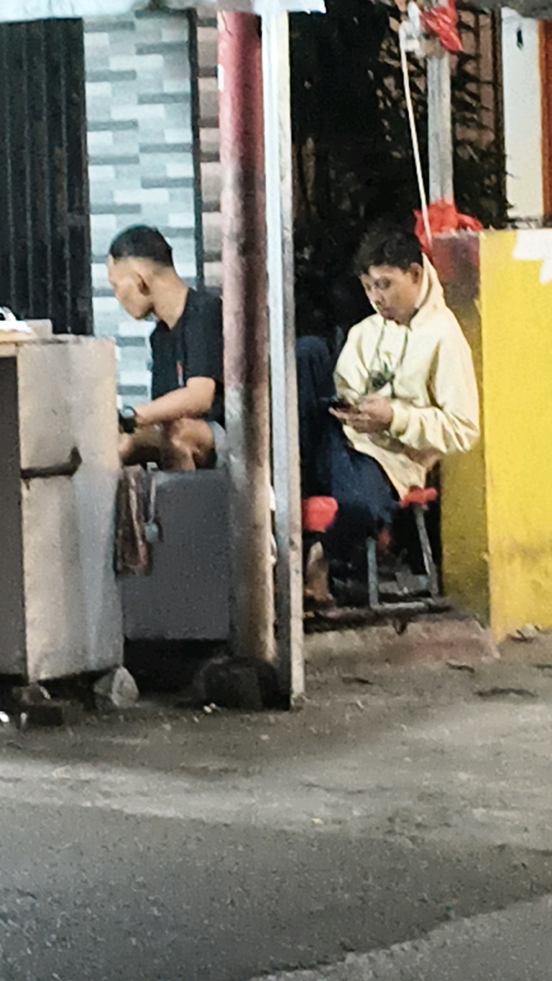 In Bekasi, Indonesia, two individuals are observed in an outdoor setting, likely a street vendor stall or open-air shop. On the left, a person wearing a black t-shirt is seated, facing away from the camera and slightly to the left. This individual appears to be interacting with an object on a counter in front of them. To the right, another person is seated on a red-seated stool, wearing a light-colored hooded jacket and dark pants. This individual is engrossed in their mobile phone, holding it in their hands and looking down at the screen. The background consists of a wall with a brick pattern on the left and a bright yellow wall on the right. A red pole stands between the two individuals. The ground is paved, with visible debris and a slight sheen, suggesting it may be wet or reflective. The overall lighting suggests it is either evening or nighttime.
