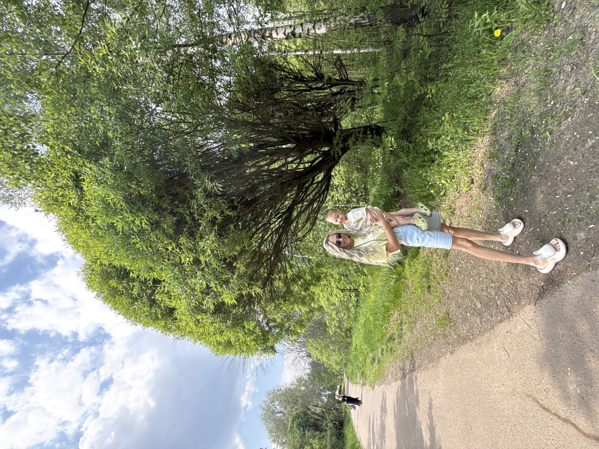 A woman with long blonde hair and sunglasses holds a young child wearing a light-colored dress and green shoes. They stand on a paved path in what appears to be a park in Rusanovo, Russia. A large, leafy tree dominates the left side of the frame, partially obscuring the sky. In the background, a figure walks along the path. The overall environment suggests a sunny, green, outdoor setting.
