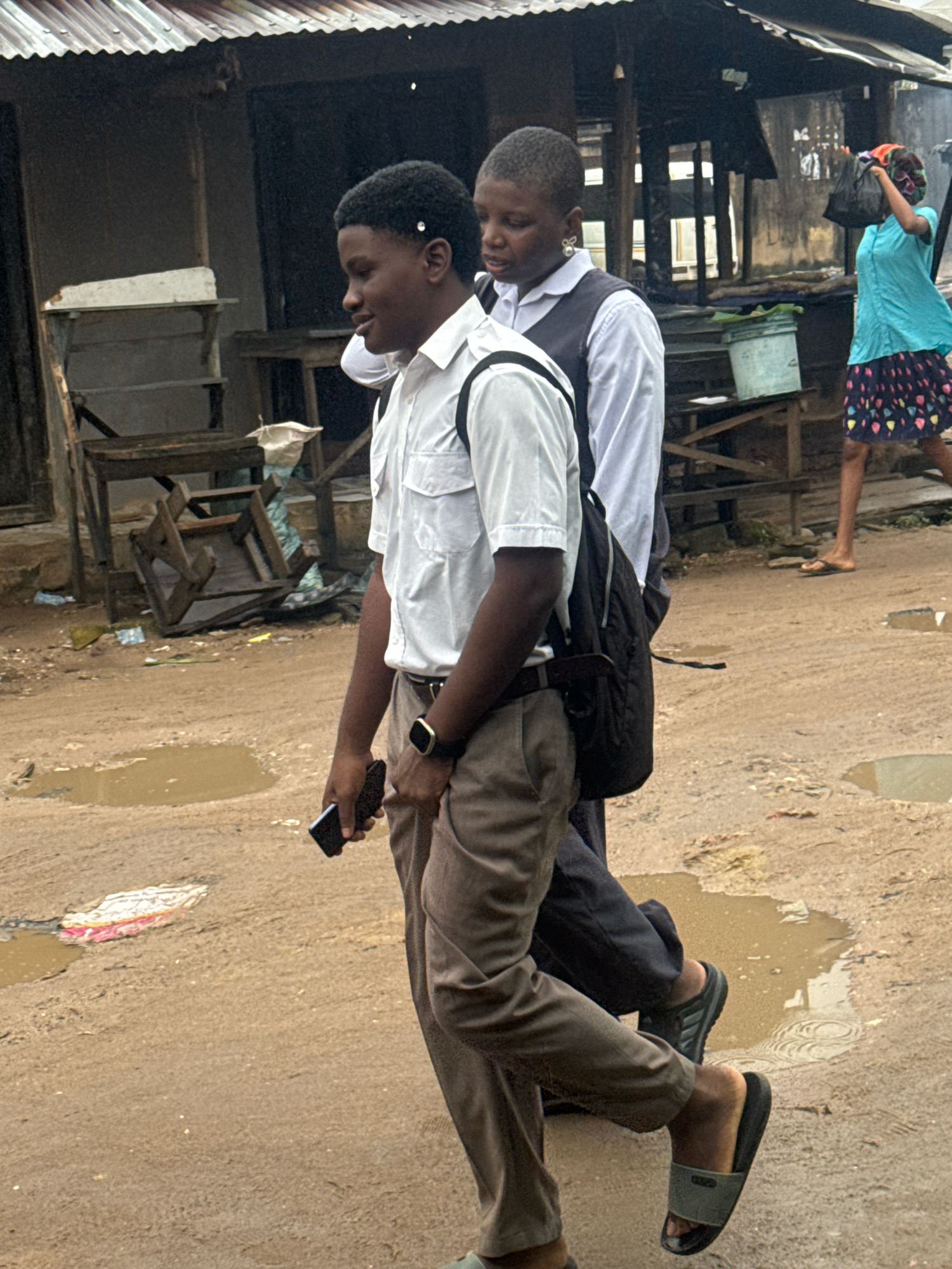 Two individuals, appearing to be students, are walking in the city of Ihiagwa, Nigeria. The person in the foreground, on the left, is a young male wearing a white short-sleeved shirt, brown trousers, and black slides. He is carrying a black backpack and holding a black smartphone in his right hand. He is looking forward and to the left. Beside him and slightly behind, on the right, is another individual, possibly a young female, wearing a white long-sleeved shirt and a dark vest. They are also looking forward. The ground is a dirt path with several puddles, indicating recent rain or damp conditions. In the background, there are structures that suggest a market or informal settlement, with corrugated metal roofs and wooden shelving units. A woman in a blue shirt and patterned skirt is visible in the middle distance, carrying a black bag over her shoulder and walking away from the camera. The scene depicts an outdoor environment in a potentially underprivileged area of Ihiagwa.