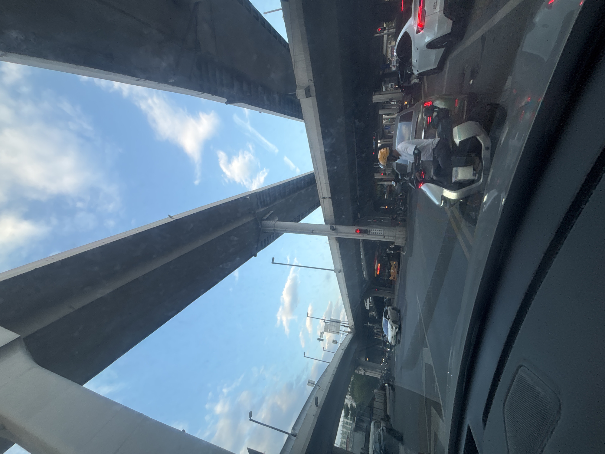 This image captures a street-level view under an elevated highway interchange in Liwan District, China. The perspective is from inside a vehicle, looking forward. Multiple concrete highway ramps dominate the upper portion of the frame, creating a geometric ceiling against a partly cloudy blue sky. Below, traffic flows. Several cars and a bus are visible on the road. A person wearing a yellow helmet rides a futuristic-looking electric scooter. The scene appears to be at an intersection, with traffic lights visible, one of which is red. The overall impression is of a busy urban environment characterized by significant infrastructure and vehicular movement.