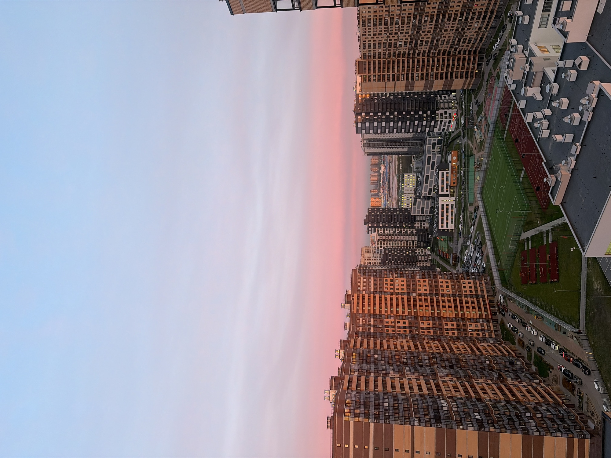 The image is an aerial view of a cityscape at sunset, likely in Saint Petersburg, Russia, based on the architecture. The sky is a gradient of pink and blue. The city is comprised of dense, high-rise apartment buildings. Between the buildings, there are green spaces including a soccer field. There are also several rows of parked cars. The photo appears to have been taken from a high vantage point, possibly from another tall building. The lighting suggests that it is either sunrise or sunset.