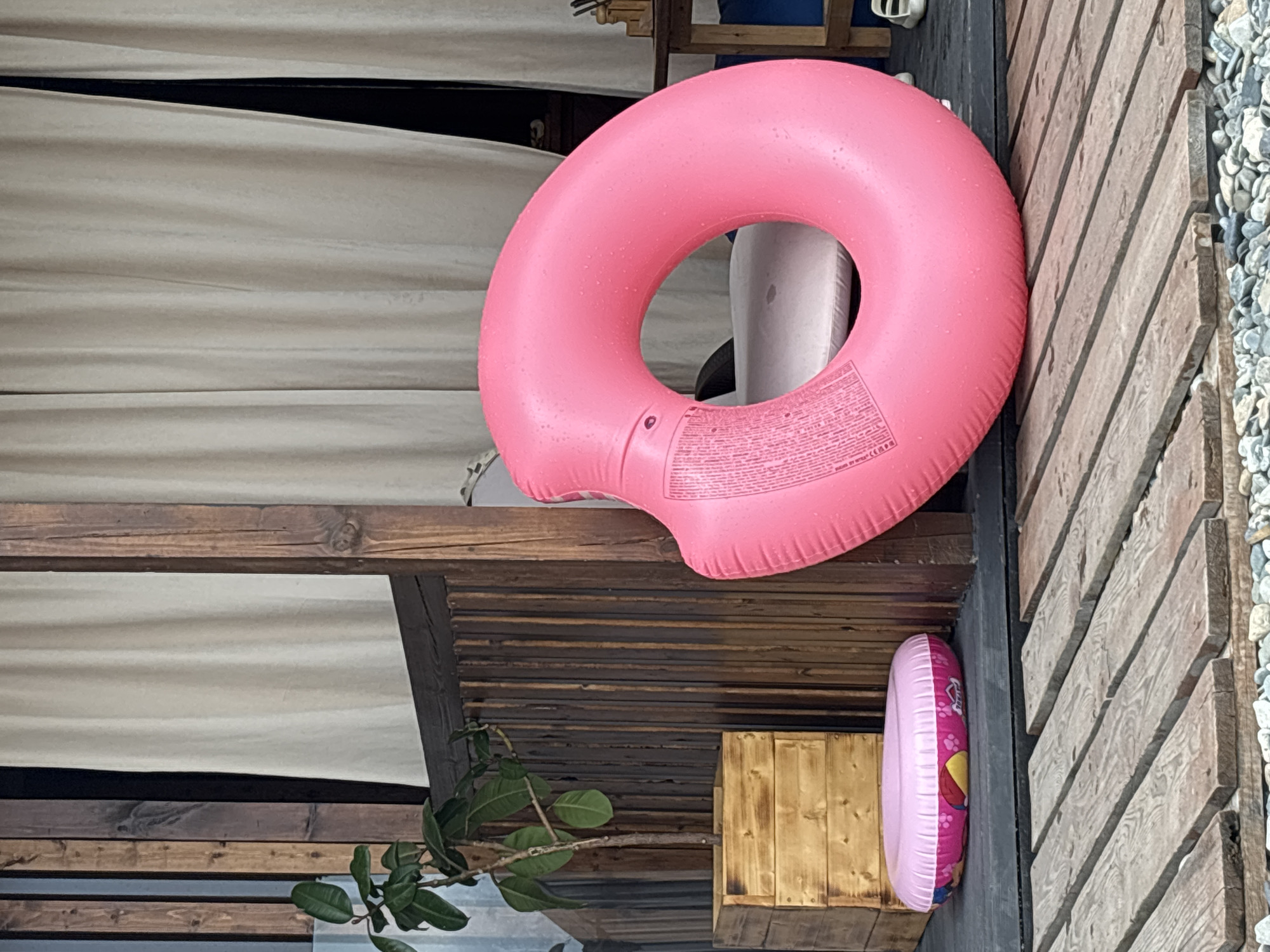 This image depicts an outdoor scene, possibly a patio or deck area, adjacent to a structure with wooden vertical siding and cream-colored curtains. In the foreground, a wooden plank walkway is visible, with a bed of pebbles at its base. A large, bright pink inflatable ring is the most prominent object, positioned centrally and angled to the right. To the left of the pink ring, a smaller, flat, pink inflatable ring with a pattern featuring characters from Paw Patrol rests on a dark gray surface. Behind the inflatable rings, a wooden planter box holds a small green plant with dark leaves. The background features more wooden siding and the draped curtains. The overall impression is one of a relaxed, possibly recreational, outdoor space. The scene is consistent with a typical summer setting in a resort town like Sochi, Russia, where outdoor living spaces are common for leisure activities.