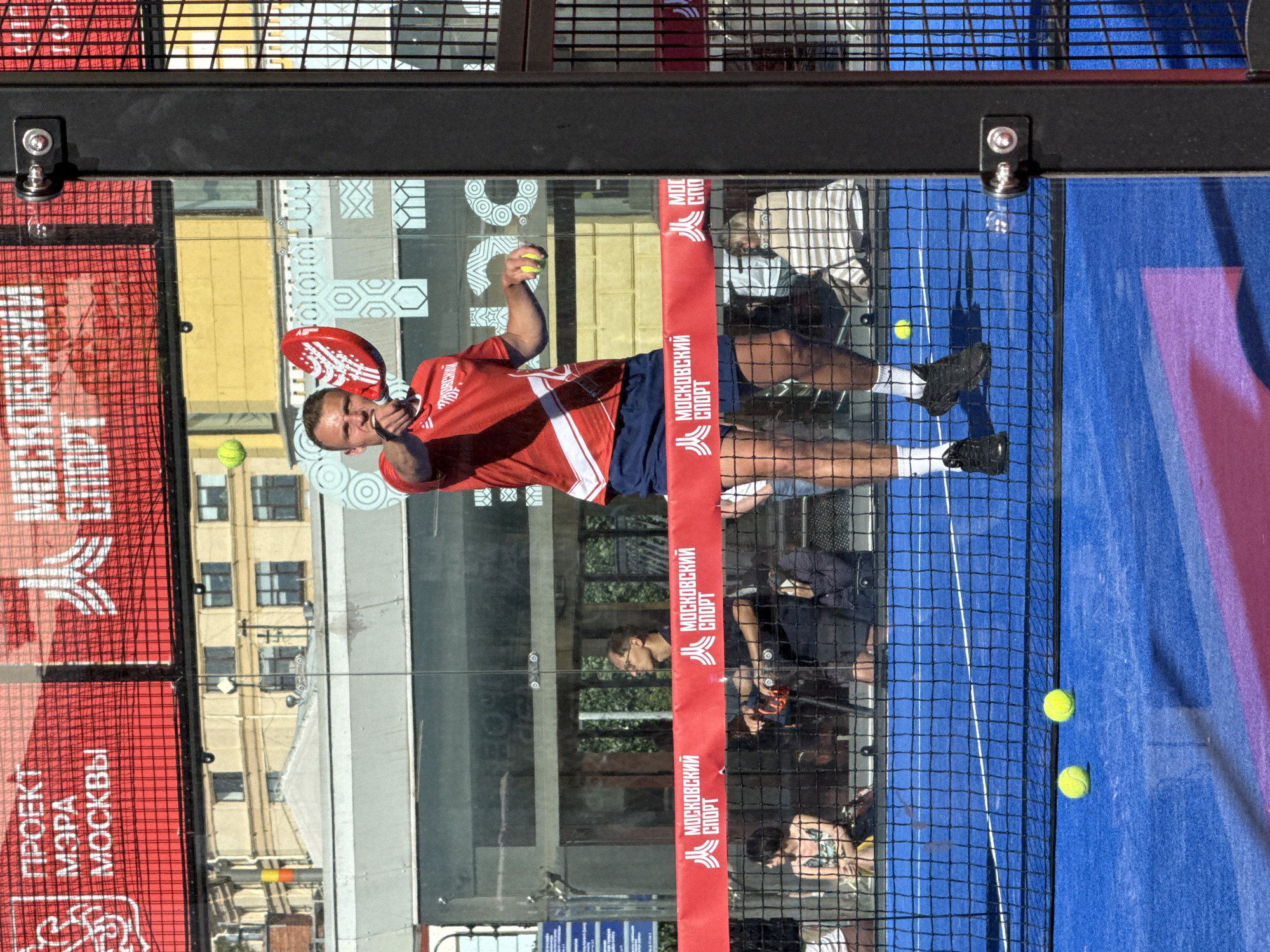 An outdoor padel tennis court in Moscow, Russia, is the setting for this image. The court is blue with a white line marking the boundaries. A black net divides the court. In the foreground, two yellow padel tennis balls rest on the blue court surface. A man in a red shirt and blue shorts is playing padel. He is in the middle of hitting a yellow ball with a red padel racket. Another yellow ball is visible in the air near the net. Spectators are visible in the background, seated behind the court. A large red banner with white text is visible above the spectators, displaying "ПРОЕКТ МЭРА МОСКВЫ" (Project of the Mayor of Moscow) and "МОСКОВСКИЙ СПОРТ" (Moscow Sport). The scene appears to be a sunny day, with the sun casting shadows.