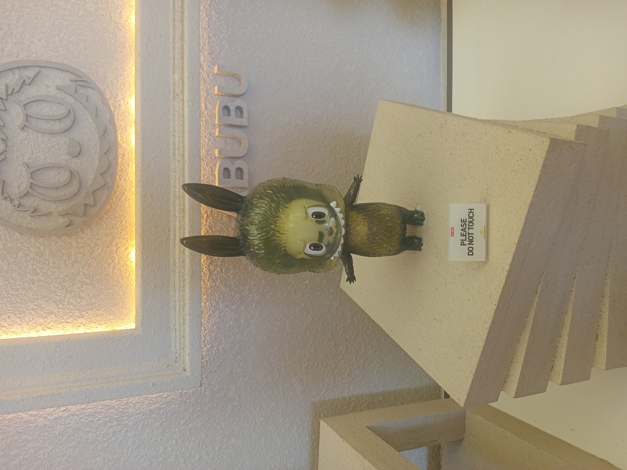 A small, stylized toy figure with large ears and a furry body stands on a stack of light-colored, textured blocks. The figure has a greenish-yellow face with wide eyes, sharp teeth, and a tuft of dark green hair. A white sign with black text reading "PLEASE DO NOT TOUCH" is affixed to the front of the blocks. In the background, the word "BUBU" is embossed on a textured wall, and a circular relief of a face with large eyes is visible above. The environment appears to be indoors, possibly a display in a store or exhibition, with soft lighting. The location is specified as Đà Nẵng, Vietnam.