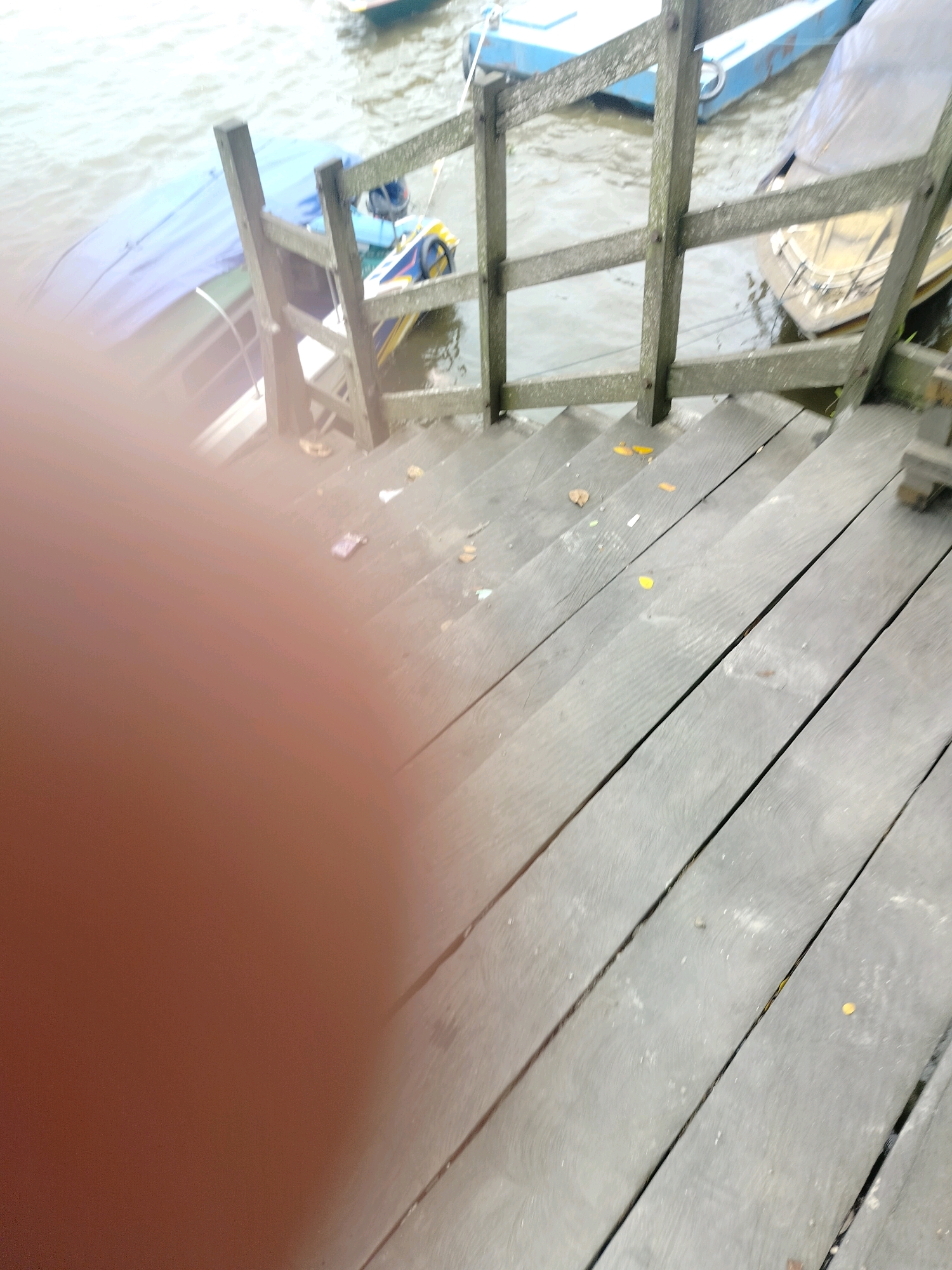 The image shows a wooden walkway with railings leading down towards a body of water. Several boats are moored near the walkway. One boat on the left is covered with a blue tarp. Another boat in the background appears to be a small passenger boat with yellow and blue accents. The walkway is made of weathered wooden planks and has some debris, including leaves and what appears to be trash, scattered on it. The water is a murky greenish-brown. The overall scene suggests a public docking area or pier. The location is identified as Kota Tanjung Selor, Indonesia, a city located on the Kayan River in the province of North Kalimantan.