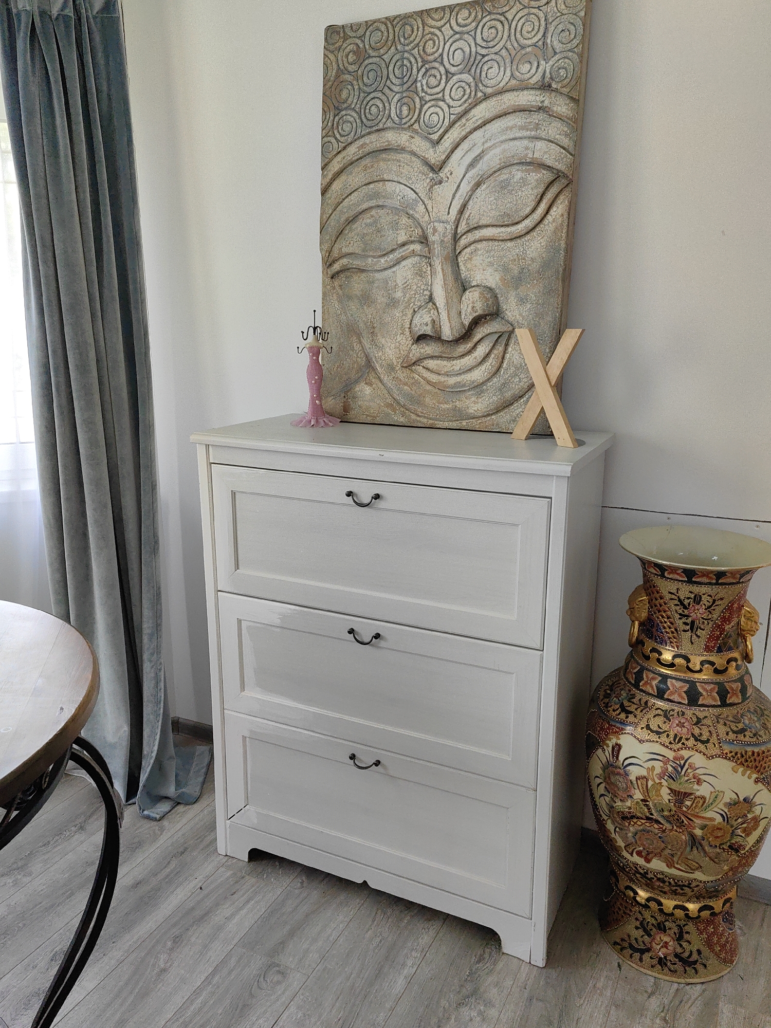 The image depicts an indoor scene, likely a room in a home, with a collection of decorative objects. In the center, a white three-drawer dresser is prominent. On top of the dresser, a large, distressed wooden wall art featuring a Buddha's face is leaning against the wall. To the right of the dresser, a tall, ornate vase with intricate floral and animal patterns in vibrant colors is placed on the floor. A small, pink, dress-shaped jewelry stand sits on the dresser, to the left of the Buddha art. A wooden letter "X" is propped up on the dresser, near the Buddha art. To the left of the dresser, a grey curtain hangs near a window, and a portion of a round wooden table with a dark metal base is visible. The flooring appears to be light-colored laminate. The lighting suggests it is daytime, with natural light coming from the left. The overall impression is one of a curated collection of decorative items in a domestic setting.