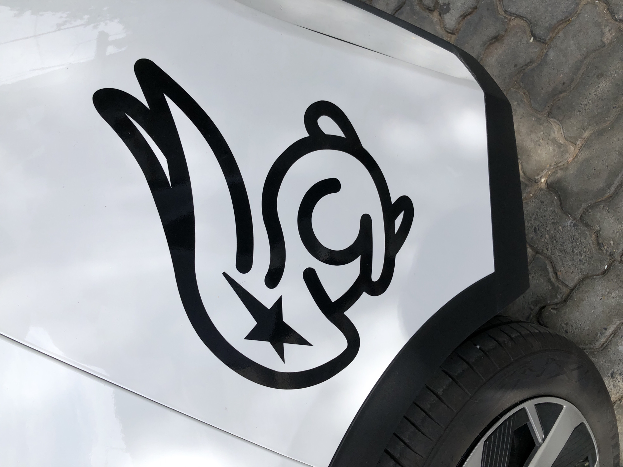 The image shows a close-up, high-angle shot of a white car fender with a black sticker of a stylized rabbit-like creature. The sticker has a star for an eye and appears to be a cartoonish design. The car is parked on a paved surface with a visible tire and black trim on the fender. The scene is outdoors, and the lighting suggests it is daytime. The image does not provide any specific geographical markers to indicate it is located in Đà Nẵng, Vietnam.