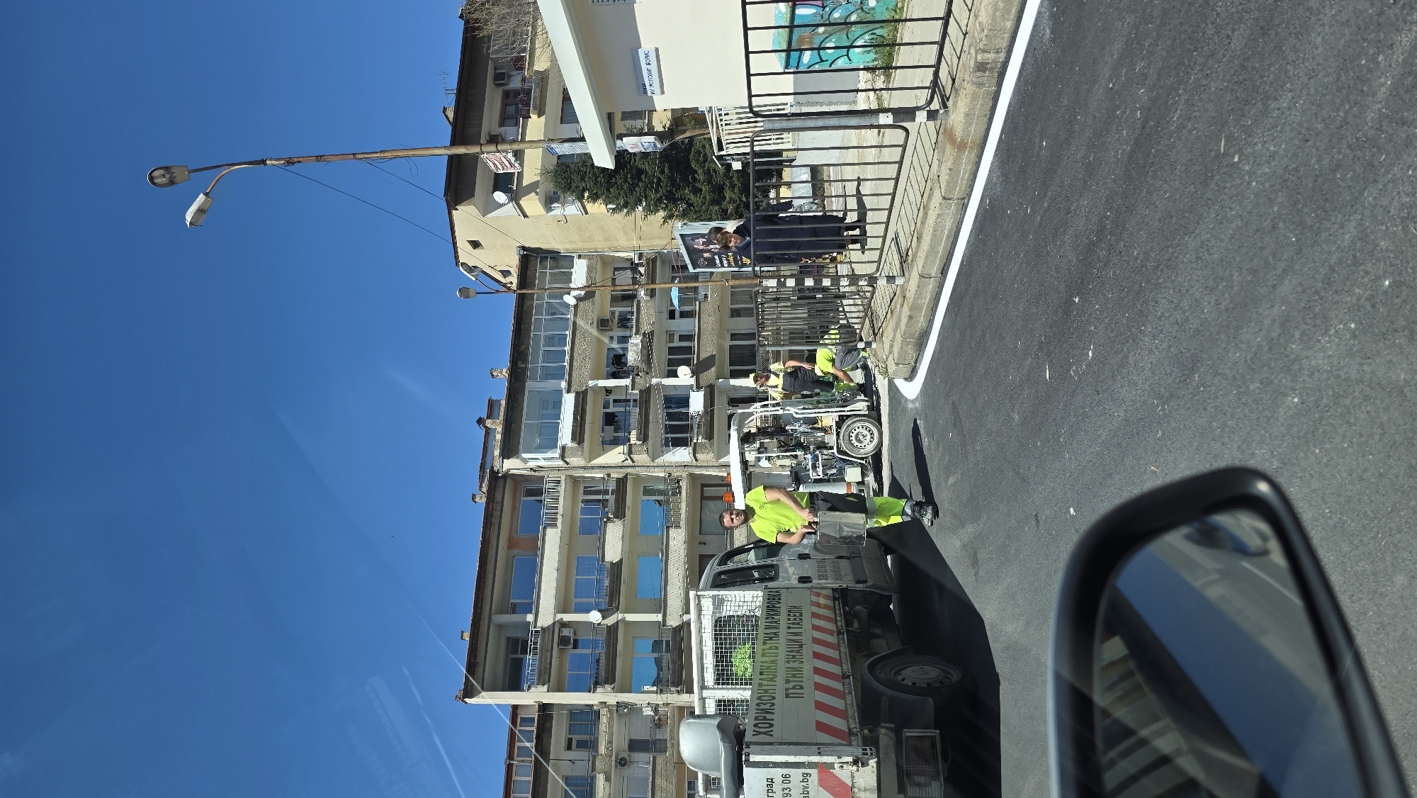 A street in Blagoevgrad, Bulgaria. A light utility truck is parked near the curb. The side of the truck has text in Bulgarian: "ХОРИЗОНТАЛНА ПЪТНА МАРКИРОВКА." Two men in lime green shirts are working on the street, one standing near the front of the truck, the other near a line marking machine. Buildings are behind the workers on the opposite side of the street. The sky is clear and blue. The image is taken from inside a vehicle, showing the side mirror.