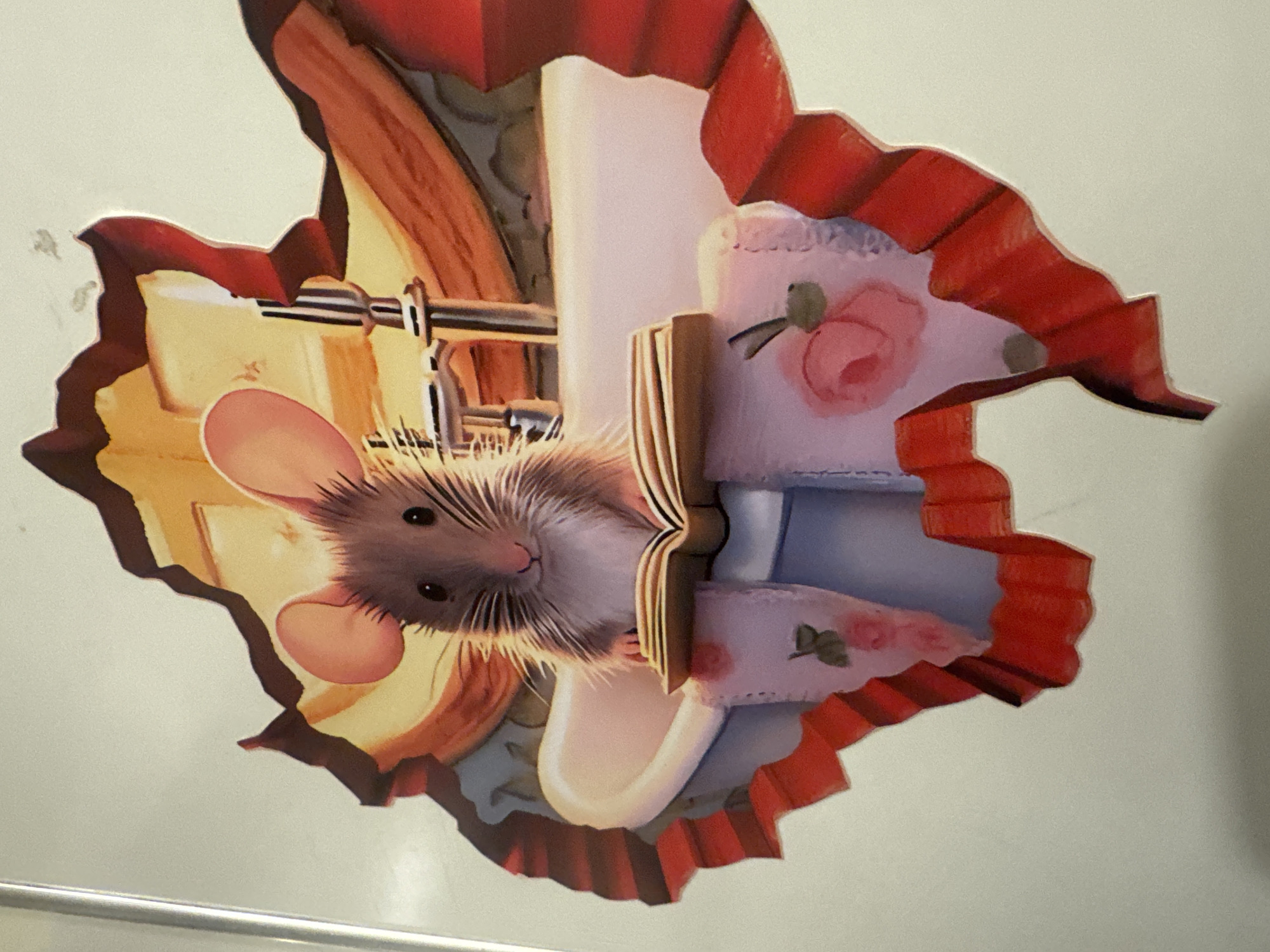 A sticker, designed to appear as if a mouse is emerging from a hole, is affixed to a wall. The sticker has a trompe-l'oeil effect, depicting a mouse with pink ears and whiskers looking out from a cut-out area, inside which appear to be pieces of furniture. The cut-out area is surrounded by a red, ruffled border, giving the impression of a hole in the wall. The sticker is located indoors. The image does not contain information about the specific location in the city of Tel-Aviv, Israel.