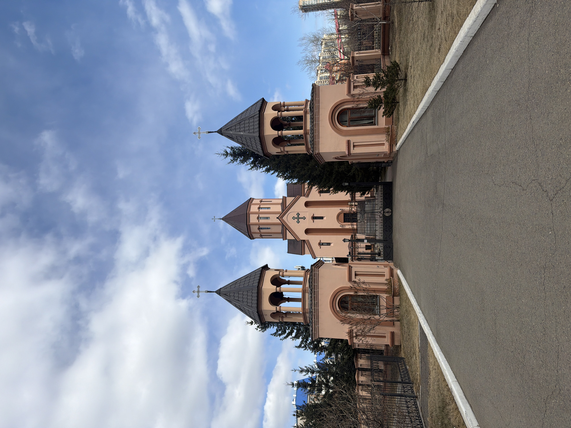 This image depicts a series of three architecturally similar church buildings, likely part of an Armenian Apostolic complex, set against a bright blue sky with scattered white clouds. The scene is captured during daylight, possibly late winter or early spring, given the bare deciduous trees.
The three main structures are light peach or pink in color, each topped with a dark grey or brown conical roof and a cross at its pinnacle.
1. The leftmost structure appears to be a bell tower, featuring an open belfry with columns supporting rounded arches.
2. The central building is larger and more prominent, also with a conical roof and a cross. It has a distinctive cross emblem positioned above its entrance.
3. The rightmost structure mirrors the design of the leftmost bell tower, complete with a conical roof and a cross.
In the foreground, a paved asphalt path with a white curb runs along the right side of the image. Beyond the curb, there's a patch of dry, yellowish-brown grass. Scattered around the church grounds and in the background are leafless trees, suggesting the seasonal timing. Ornate black metal fences enclose parts of the property. In the distant background, taller urban or suburban buildings are visible, indicating the church is situated within a developed area. The overall atmosphere is peaceful and serene, with no human activity or visible text. The location context is Krasnoyarsk, Russia.