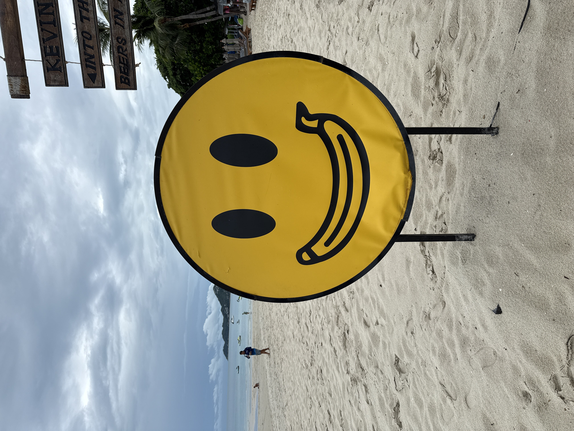 A yellow smiley face sign is in the foreground on a sandy beach in the Ko Samui District of Thailand. The sign has black eyes and a mouth shaped like a banana. In the background, there is a body of water with several boats and people on the beach. A wooden sign with carved text is hanging to the right of the smiley face. The sky is cloudy.