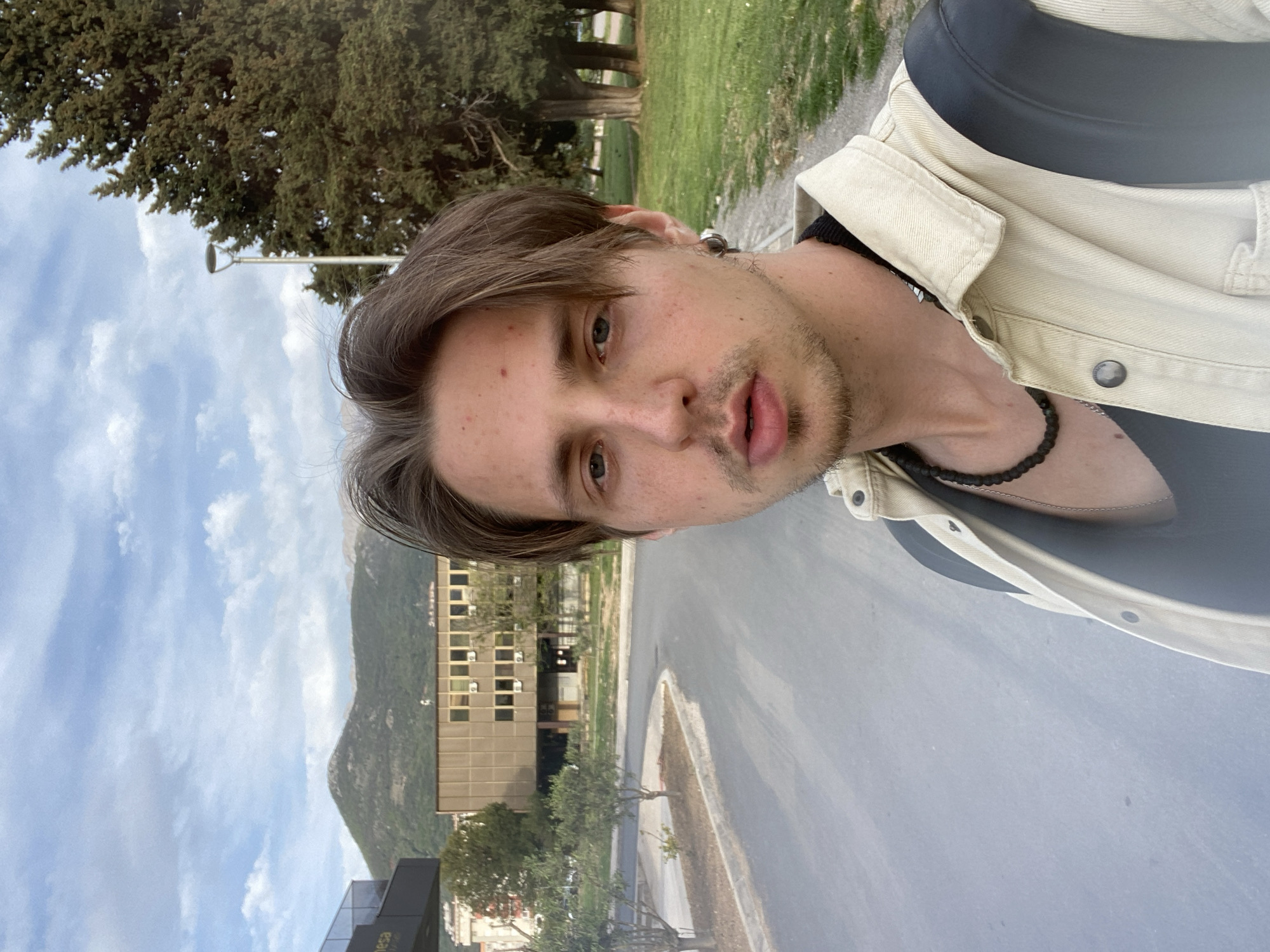 A young adult male is taking a selfie outdoors. He has light brown hair, light eyes, and stubble. He wears a black shirt under a light beige jacket and a black beaded necklace. The background consists of a paved road, a multi-story building with many windows, and a mountain in the distance under a partly cloudy sky. The location is consistent with the city of Bar, Montenegro, given the architectural style and mountainous terrain. The presence of a European-style street lamp and type of street further confirm this.