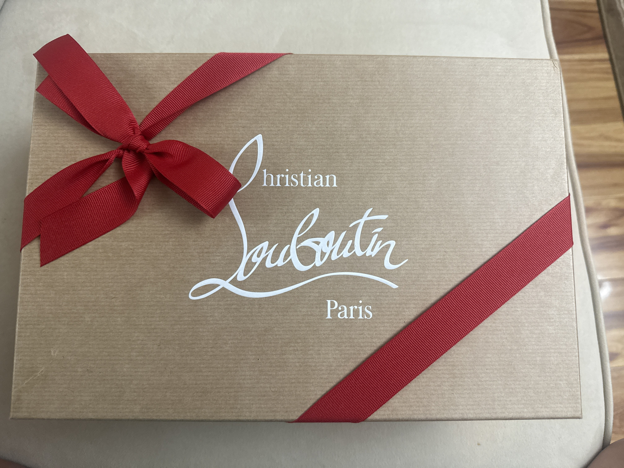 The image shows a Christian Louboutin box. The box is brown with a red ribbon diagonally across the lower right corner and tied in a bow in the upper left corner. The Christian Louboutin logo and "Paris" are printed in white on the box. The location is Hallandale Beach, United States of America, but this is not ascertainable from the image. There are no people or actions visible.