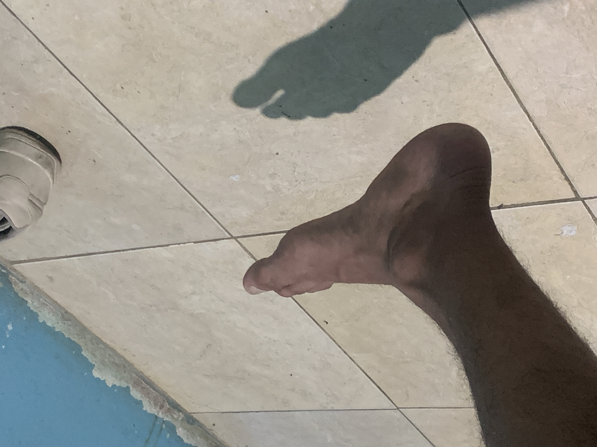 An overhead shot captures a dark-skinned foot and lower leg, along with a partial view of a white shoe, on a light-colored tiled floor. The foot, positioned prominently in the lower right, appears bare, with individual toes visible. A distinct, dark shadow of a foot, complete with separated toes, is cast on the tiles in the upper-middle section of the image, suggesting bright, possibly overhead, lighting. The tiled floor features a grid pattern with dark grout lines, and some faint marks or debris are scattered across its surface. In the lower left corner, a portion of a bright blue surface, possibly a low wall or the edge of a pool, is visible, with an uneven, white-ish border where it meets the tiled floor. The scene suggests an indoor or covered outdoor area, likely during the daytime due to the clear shadow. The location context is Santa Ana, Venezuela. There is no discernible activity beyond standing or resting, and no text is visible.