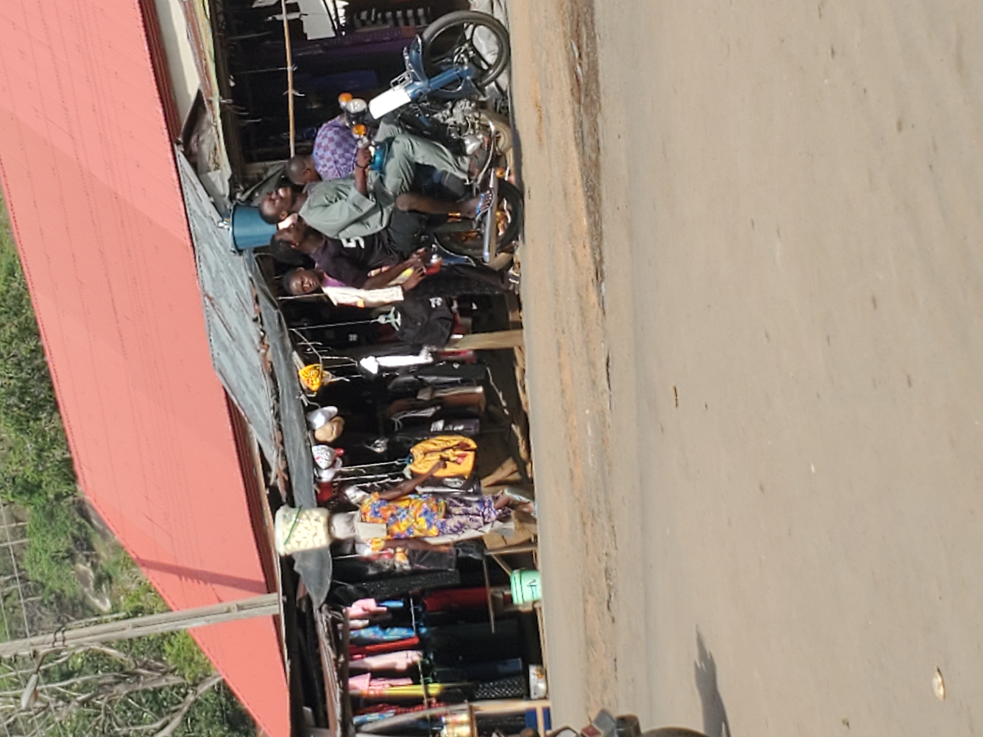 The image depicts a street scene in Mararaba, Nigeria. A row of makeshift shops lines the side of the road. The shops display a variety of merchandise, including clothes, shoes, and accessories. Several people are present, including shopkeepers and customers. A motorcycle is parked near one of the shops. The road is paved. The lighting suggests it is daytime.