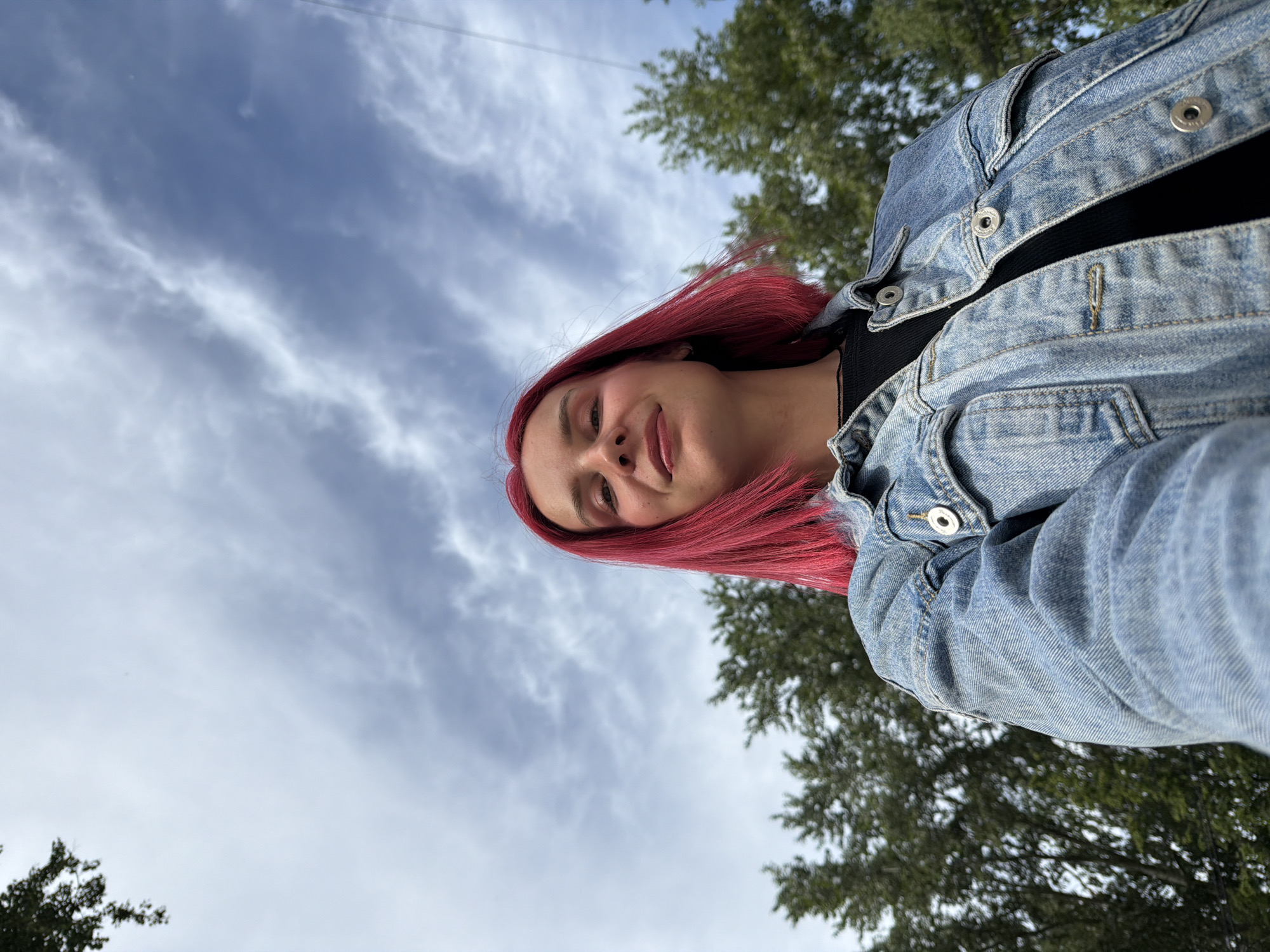 This low-angle outdoor selfie, taken in Chaikovsky, Russia, features a young person with bright red hair. They are positioned on the right side of the frame, looking downwards towards the camera with a slight, content smile. The individual is wearing a light-wash denim jacket over a black top.
The background is dominated by a cloudy sky, showing patches of blue and white, suggesting an overcast daytime. Green tree foliage is visible in the upper right and lower left portions of the image, indicating an outdoor setting, likely a park or a tree-lined area. The image is oriented with a significant counter-clockwise rotation, giving it a dynamic, off-kilter perspective. No visible text is present in the image.