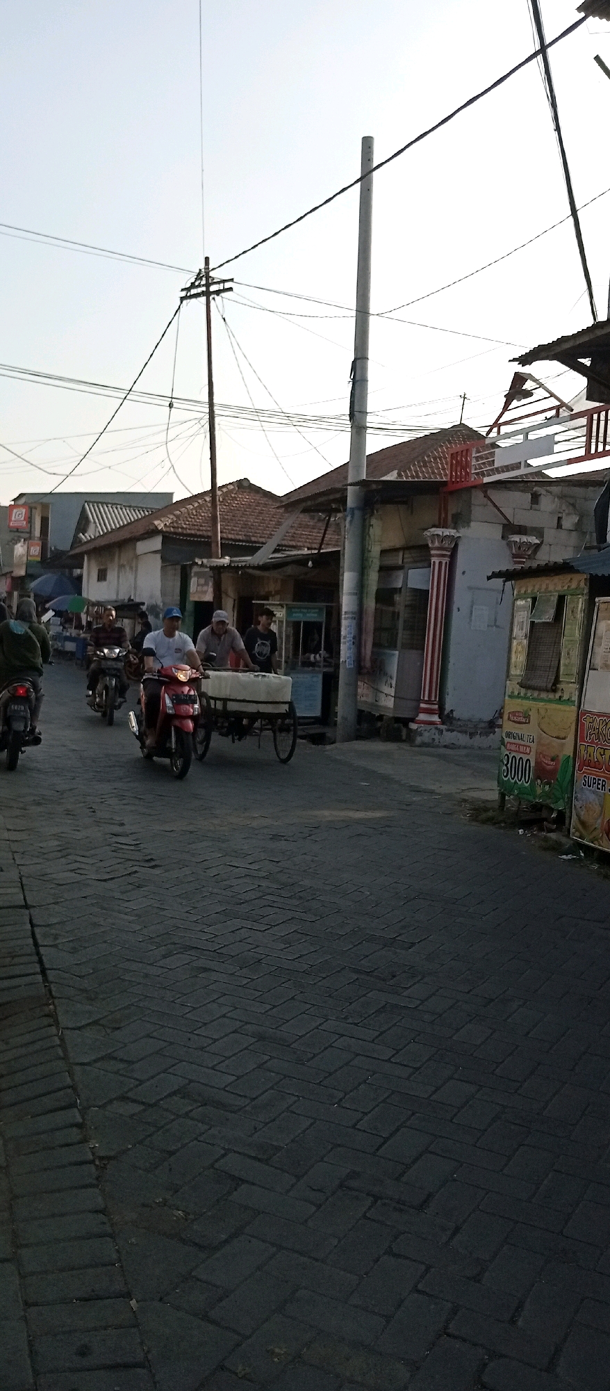 This is a low-angle, full shot of a street scene in Wadungasri, Indonesia, during the daytime, likely late afternoon given the lighting. The street is paved with dark interlocking bricks arranged in a herringbone pattern. On the right side of the street, there are several small storefronts and kiosks. One kiosk has a prominent sign displaying "ORIGINAL TEA" and the price "3000". Another kiosk has a sign that reads "TAKO JASU SUPER J". A tall, slender utility pole with multiple wires stretches across the frame. Several people are present. In the foreground, a red motorcycle is being ridden by a man wearing a light-colored cap and a grey shirt. He is pulling a cart with a white tarp covering its contents. Behind him, another motorcycle is visible. Further down the street, several other motorcycles and people are seen, suggesting moderate activity. Buildings with tiled roofs line the street on the left and right. The sky is clear and pale.