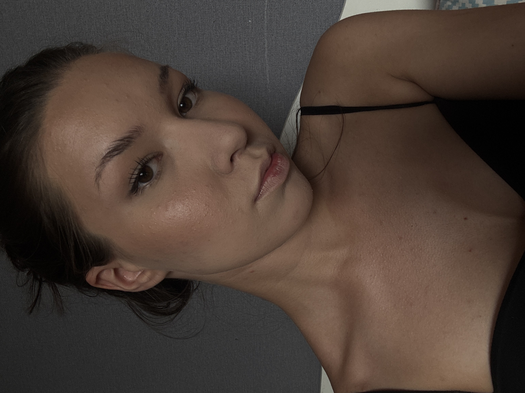 The image is a close-up, head-and-shoulders portrait of a young woman with brown hair pulled back and brown eyes. She is wearing a black camisole. Her skin appears to have makeup applied, with blush on her cheeks and mascara on her eyelashes. The background is a textured gray surface. There are no identifiable elements that place the image in Vishnevka, Russia or any other specific location.