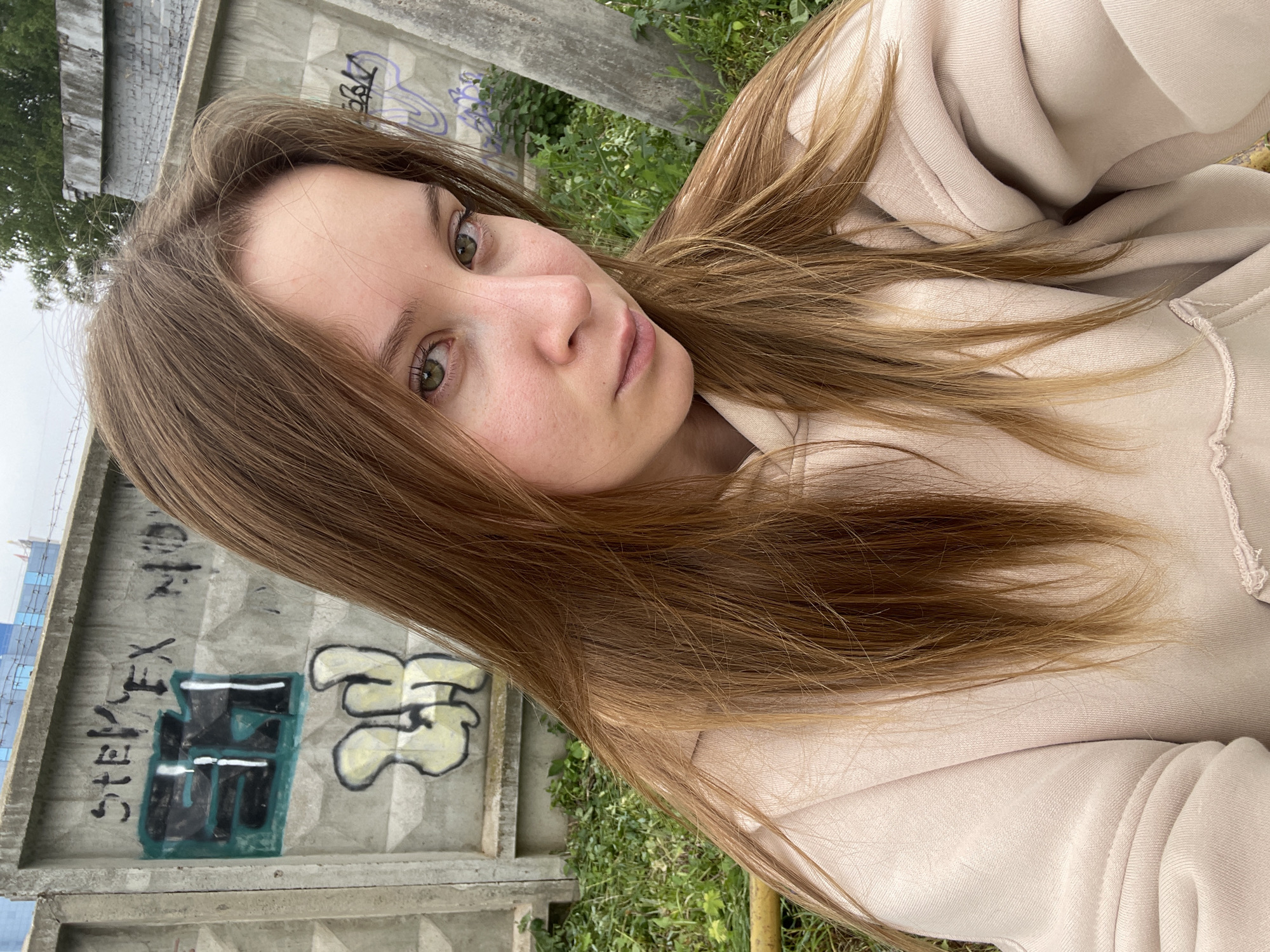 In Chaikovsky, Russia, a young woman with long, light brown hair is photographed from the chest up. She is wearing a light beige hoodie. Her gaze is directed towards the camera, and her expression is neutral. Behind her, a concrete fence is visible, adorned with various graffiti tags. The fence appears weathered and is set against a backdrop of green foliage and a cloudy sky. A portion of a yellow railing is also visible in the lower left quadrant of the image.