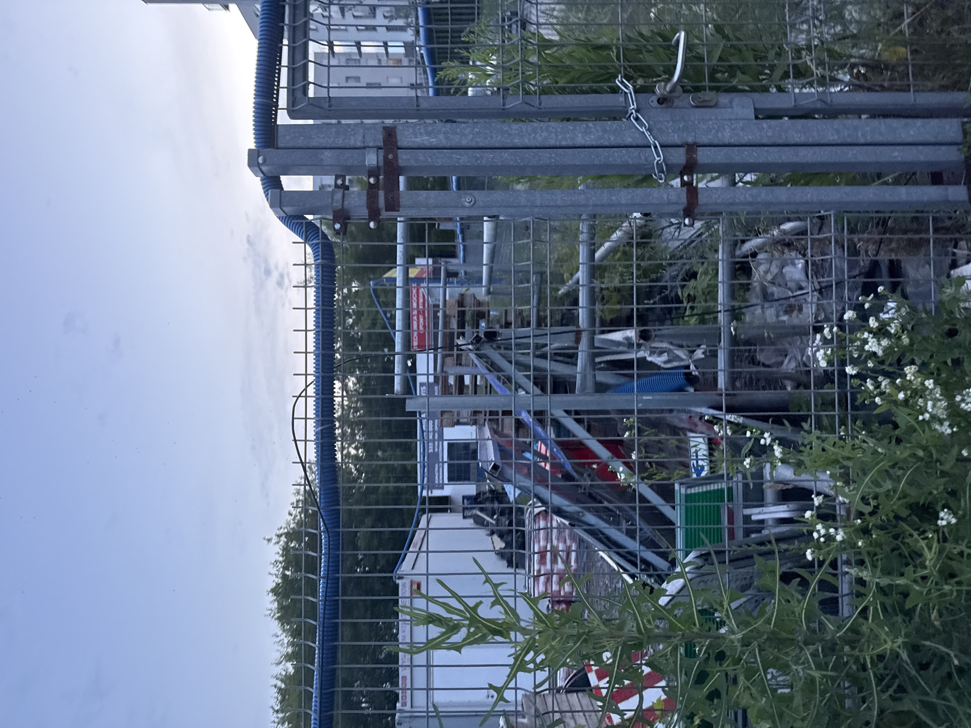A chain-link fence gate is partially open and secured with a chain and padlock. A silver handle is visible on the right gatepost. Behind the fence, construction materials are stored, including portable buildings, pallets, scaffolding, and various debris. Vegetation, including tall green plants with white flowers, grows in the foreground. The sky above is overcast.