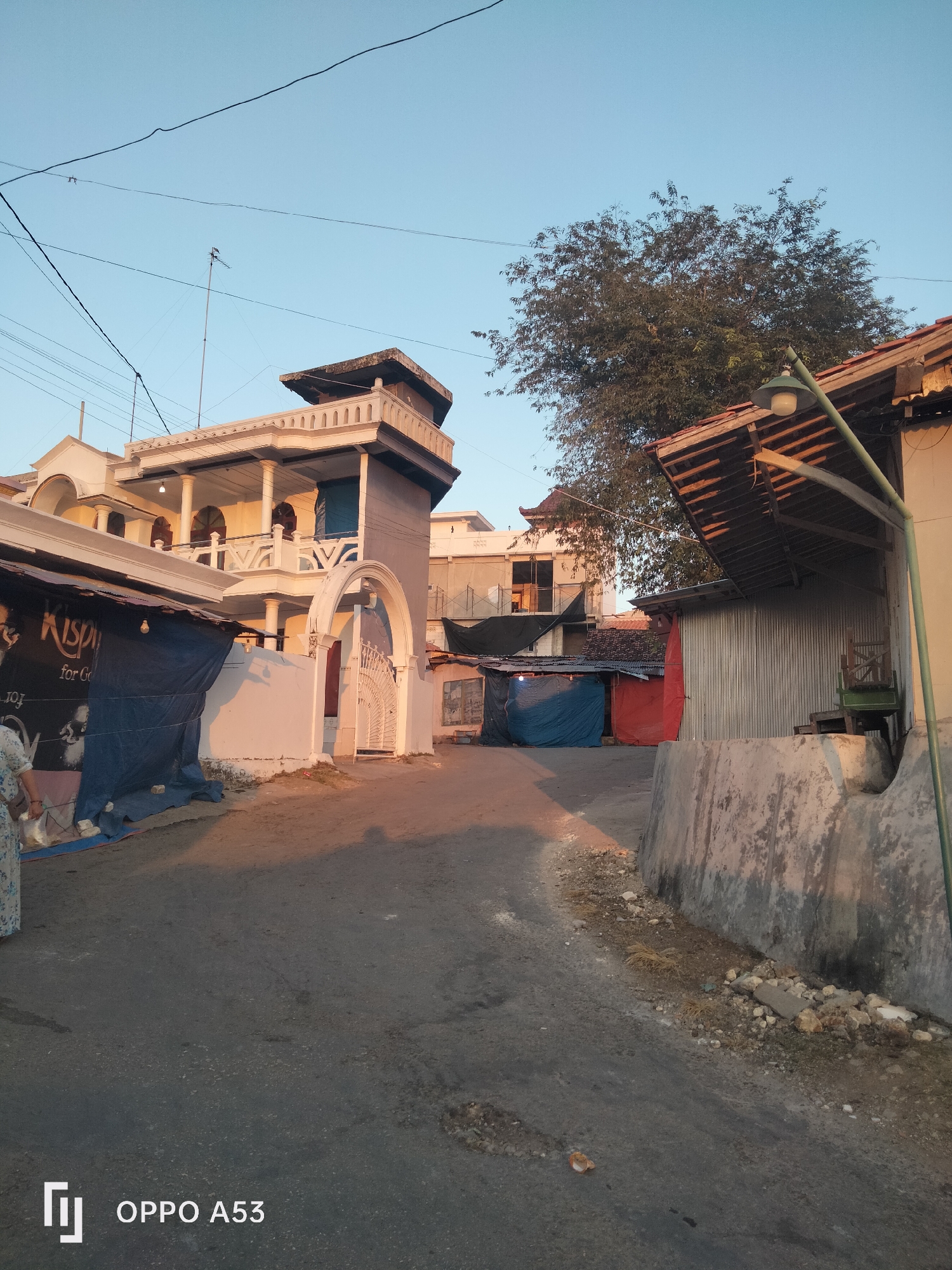 The image depicts a street scene at what appears to be dawn or dusk in Badung, Indonesia. A narrow asphalt road runs through the scene, with buildings on either side. On the left, a multi-story building with white ornate balconies and arched entrances is prominent. It has a modern yet somewhat colonial architectural style. Adjacent to it, a lower structure covered with blue tarpaulin is visible, with a sign that appears to read "Krispi for God." A person, partially visible on the far left, is wearing a patterned dress and carrying a bag.
On the right side of the street, a building with a corrugated metal facade is partially obscured by a sloped, tiled roof. A street lamp with a green pole and a white bulb is attached to this structure. Further back, other buildings with various roofing materials and some construction materials are visible. A large tree with dense foliage stands behind the buildings on the right. The sky is a clear, pale blue, indicating good weather. The lighting suggests a low sun angle, casting long shadows across the road and walls, and bathing the scene in a warm, soft glow. The overall impression is of a residential or mixed-use urban area in Indonesia.