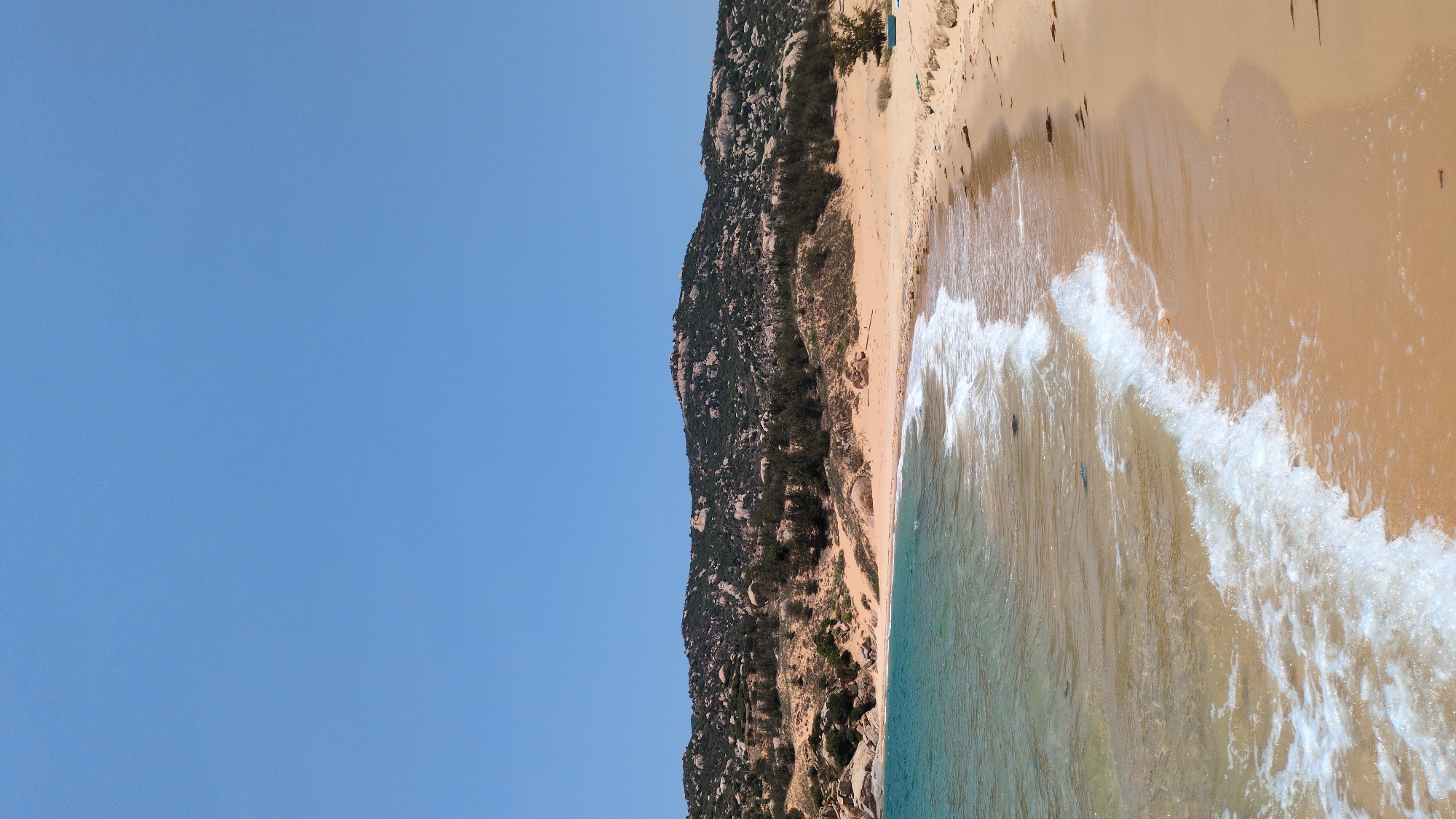 The image depicts a coastal scene in Phuoc Dinh, Vietnam. A clear blue sky dominates the left side of the frame. A rocky, vegetation-covered hill runs along the upper central part of the image. To the right of the hill, a sandy beach stretches toward the coastline. Waves break along the shoreline.
