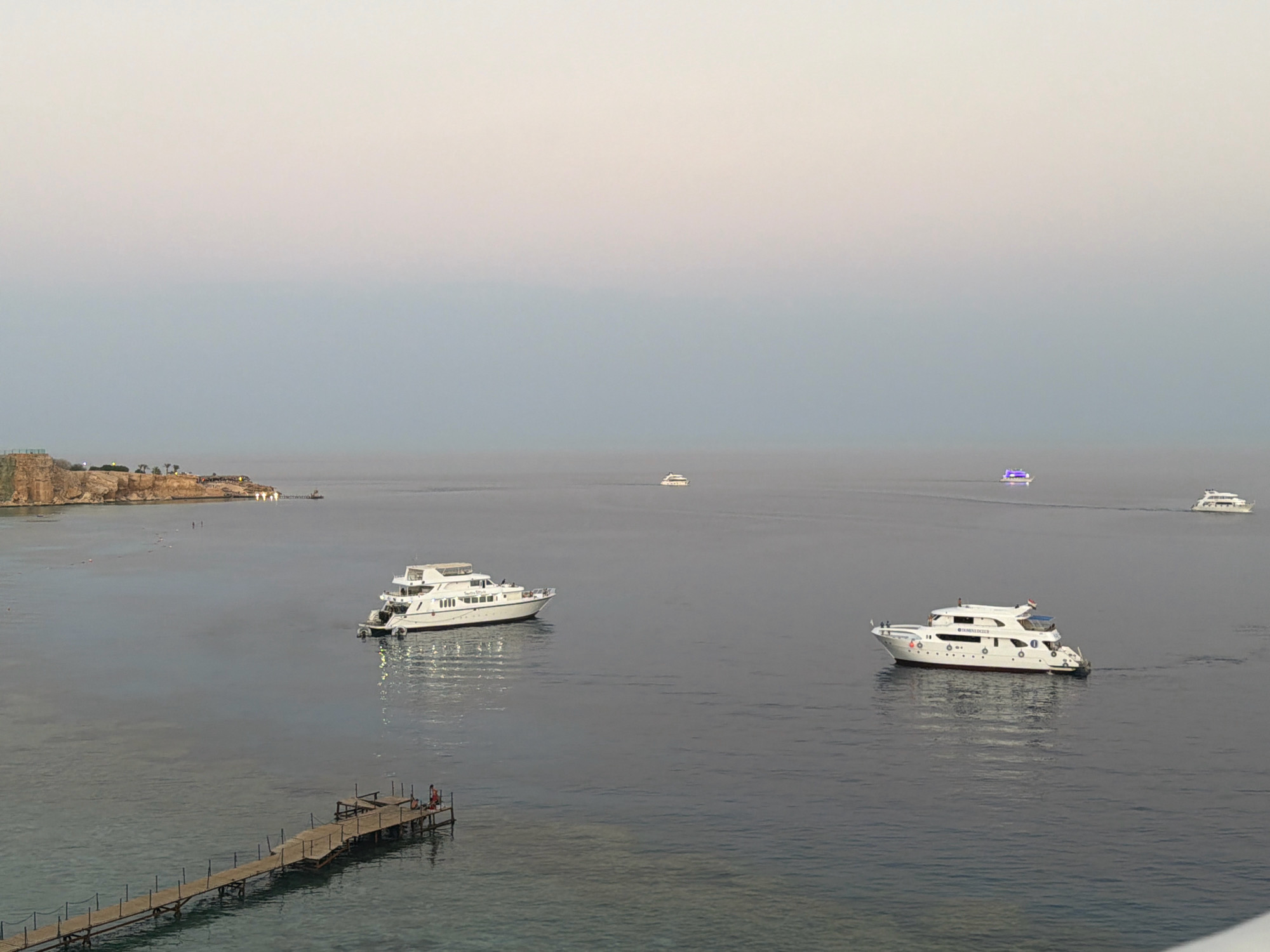 The image captures a serene seascape at dusk in Sharm El Sheikh, Egypt. In the foreground, a wooden pier extends into the calm, dark blue water. Two individuals, appearing to be men, are visible on the pier, one standing and one seated.
Further out in the water, a large white yacht is docked. Several other boats, including a smaller white yacht and a boat with colorful lights, are visible in the distance. A rocky coastline with buildings and lights is situated on the left side of the frame, likely representing part of the city's shoreline. The sky is a gradient of pale pink and blue, indicating the fading light of day. The overall atmosphere is tranquil and peaceful, showcasing the coastal beauty of Sharm El Sheikh.