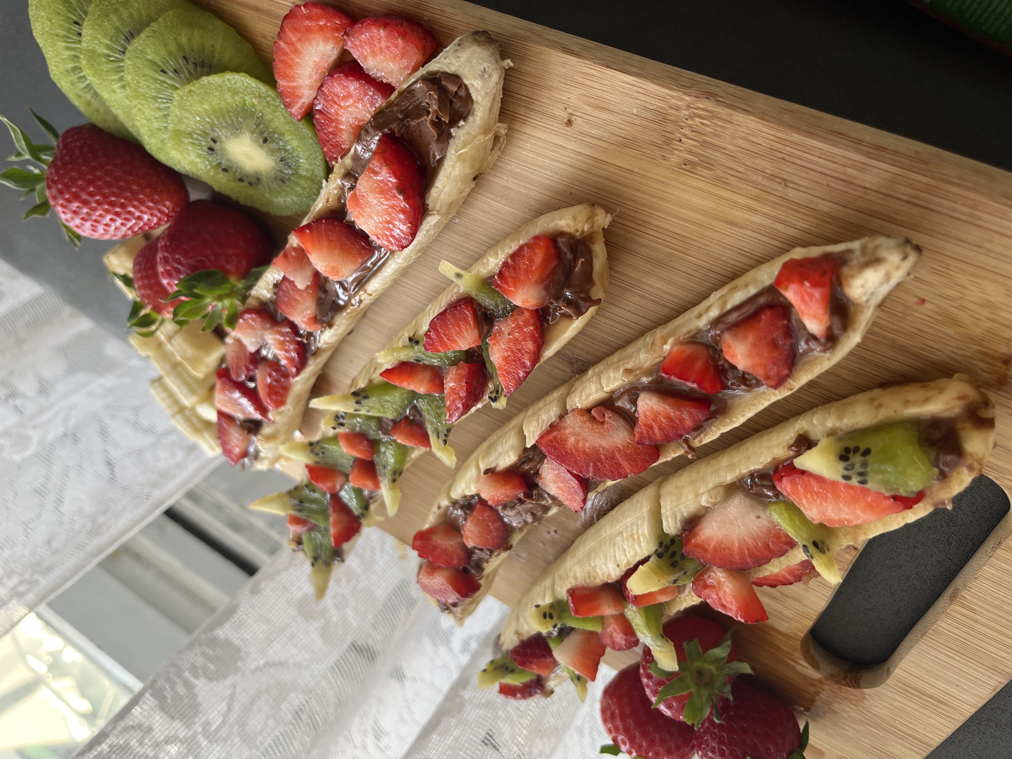The image displays a vibrant, sweet snack or dessert platter arranged on a light-colored wooden serving board. The primary subjects are several "banana boats," created by splitting bananas lengthwise and filling them with a rich, dark spread, likely chocolate or Nutella. These are generously topped with diced red strawberries and bright green kiwi pieces. Four such banana boats are visible, arranged diagonally across the board.
To the upper left, fresh ingredients are presented: several whole, ripe red strawberries with their green stems, and neatly sliced rounds of green kiwi, showcasing their characteristic black seeds. Additional diced strawberries and kiwi are scattered around the lower banana boats, adding to the colorful and appealing presentation.
The scene appears to be set indoors during daytime, illuminated by bright, natural light from the left. A white, textured fabric, possibly a lace tablecloth or curtain, is visible in the background on the left and bottom. Below this fabric, a slightly reflective surface, perhaps a window or mirrored table, shows blurred light reflections. The upper right portion of the image is a dark, out-of-focus area. There is no clearly readable text present. The overall impression is a freshly prepared, inviting treat, possibly for breakfast or a light dessert, in Sofia, Bulgaria.