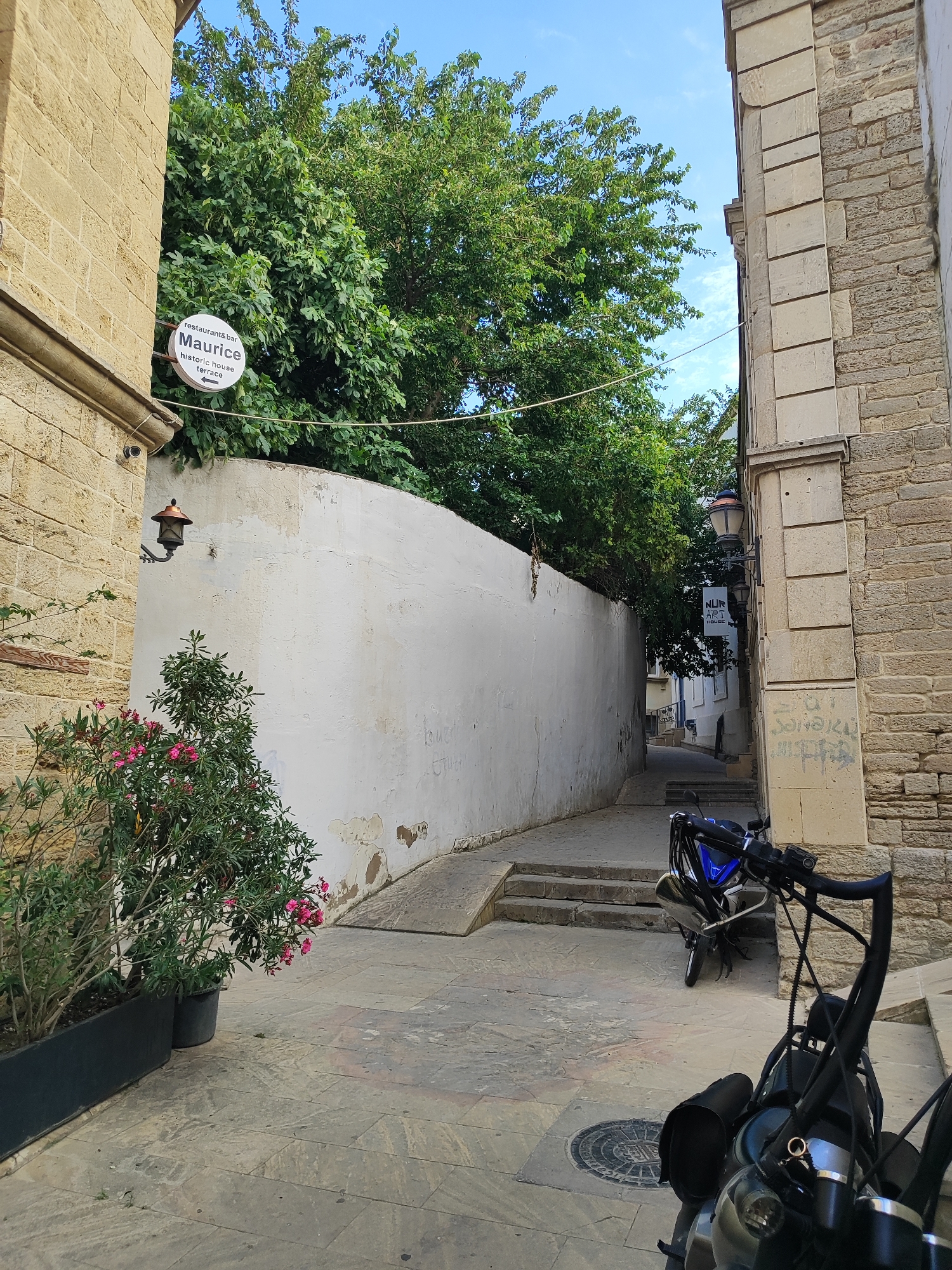 This media file captures a narrow, stone-paved alleyway flanked by buildings constructed from light-colored stone. The alley recedes into the distance, leading to more buildings and trees. The sky above is bright blue with a few wispy clouds, suggesting it is daytime with clear weather.
On the left side of the alley, a building features a circular white sign with black text that reads "restaurant & bar Maurice historic house terrace" and an arrow pointing to the left. Below the sign, a vintage-style lantern with a copper top is mounted on the wall. A potted plant with green leaves and pink flowers is situated in front of this building.
On the right side of the alley, a stone building displays some graffiti. A lamppost with two lamps is attached to this building. Further down the alley, another sign, this one rectangular and white with black text, reads "NUR ART HOUSE".
Two motorcycles are visible in the alley. One, a blue and black motorcycle, is parked further down the lane, while the handlebars and front section of another, darker motorcycle are in the foreground, suggesting the photographer is standing behind it. The overall scene depicts a tranquil, historic urban environment.