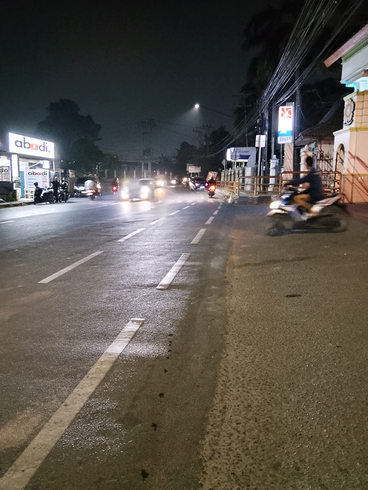 The image depicts a street scene at night in Palembang, Indonesia, likely on a road with businesses lining one side. The asphalt road is wet, reflecting the lights of passing vehicles. A prominent "abadi." sign is visible on a building to the left, with another smaller "abadi." sign below it. Several motorcycles are parked near the buildings. On the road, multiple vehicles are in motion, with their headlights and taillights creating streaks of light due to the long exposure. A person on a motorcycle is prominently captured in motion, slightly blurred, on the right side of the image. The sky is dark, with some atmospheric haze or light pollution visible. Electrical wires are strung overhead, and a street light is present in the distance. The overall impression is a busy urban street at night, with typical traffic and commercial activity.