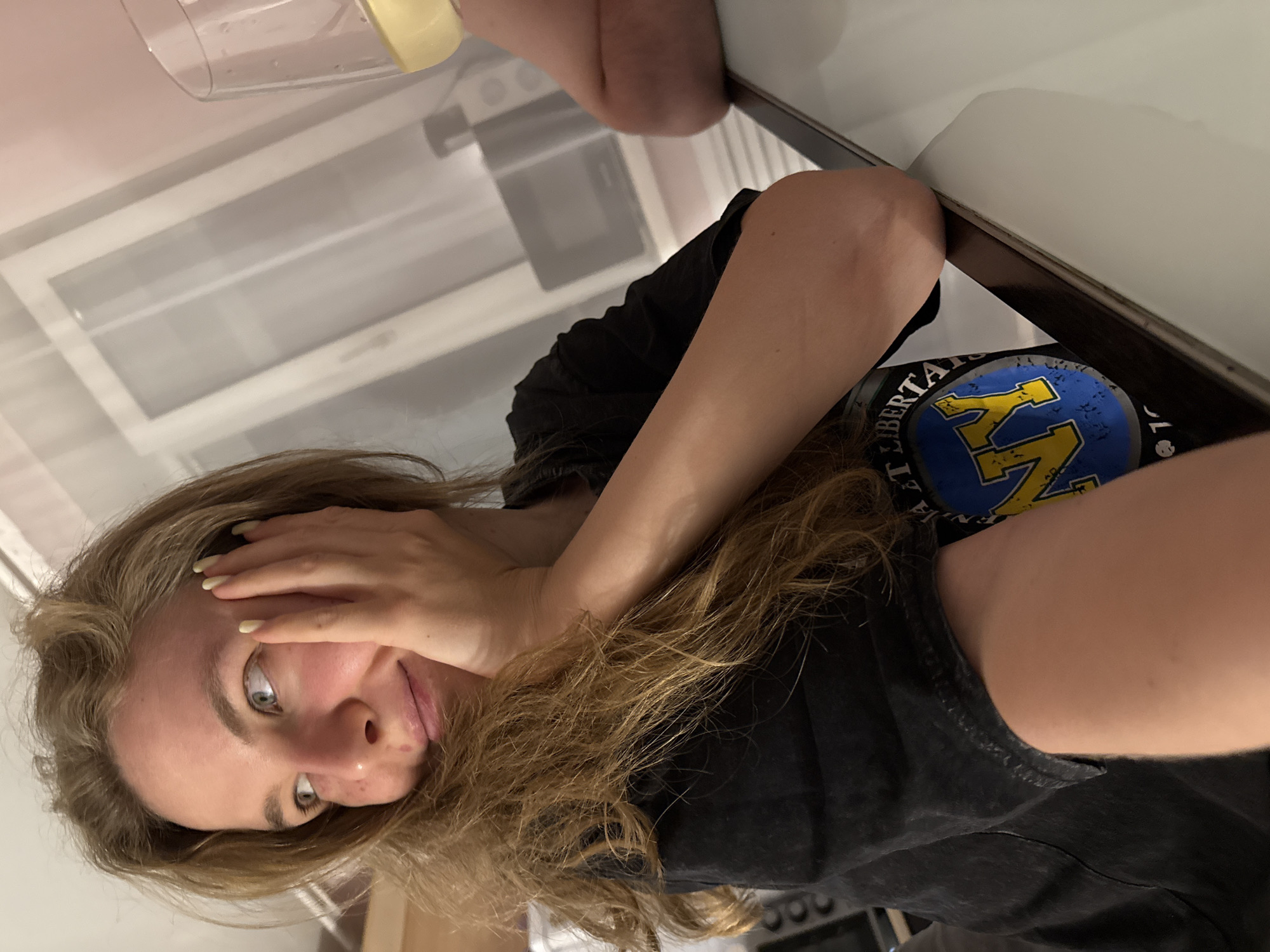 The image presents an adult female with long, wavy, light brown hair and blue-green eyes, appearing to be in her 20s or 30s. She is seated at a light-colored table, resting her left elbow on its surface and her left hand against her cheek. Her gaze is directed off-camera to her left, accompanied by a subtle smile. She wears a black, short-sleeved t-shirt with a distressed blue and yellow 'NY' logo, partially visible on her chest. Her fingernails are painted a pale yellow.
On the table to her right, a clear stemmed wine glass holds a light yellow liquid. A person's arm, presumably belonging to another individual, is partially visible behind the glass. The background features a white paneled door, a section of pale pink wall, and a silver microwave oven situated on a distant countertop. The setting appears to be an indoor domestic environment, possibly a kitchen or dining area. The image provides no discernible visual cues or identifiers to confirm its location in the city of Vladimir, Russia.