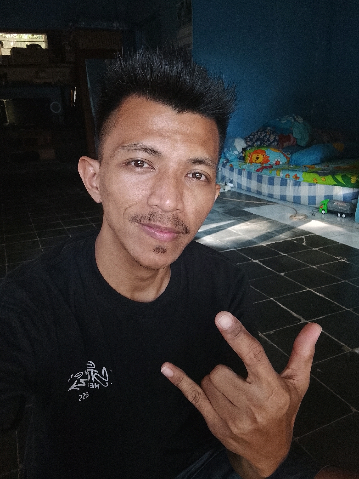 The image is a selfie taken by a man in what appears to be a bedroom or living space. The man, with dark hair styled upwards and a mustache and goatee, is wearing a black t-shirt with white graphic print. He is making a hand gesture with his right hand, forming a "rock on" sign. The background shows a bed with colorful bedding and a blue wall, suggesting a domestic environment. The flooring is tiled. The lighting indicates it might be daytime, with sunlight casting shadows on the floor. The location is identified as Kertahayu, Indonesia, though no specific landmarks are visible to confirm this within the image itself.