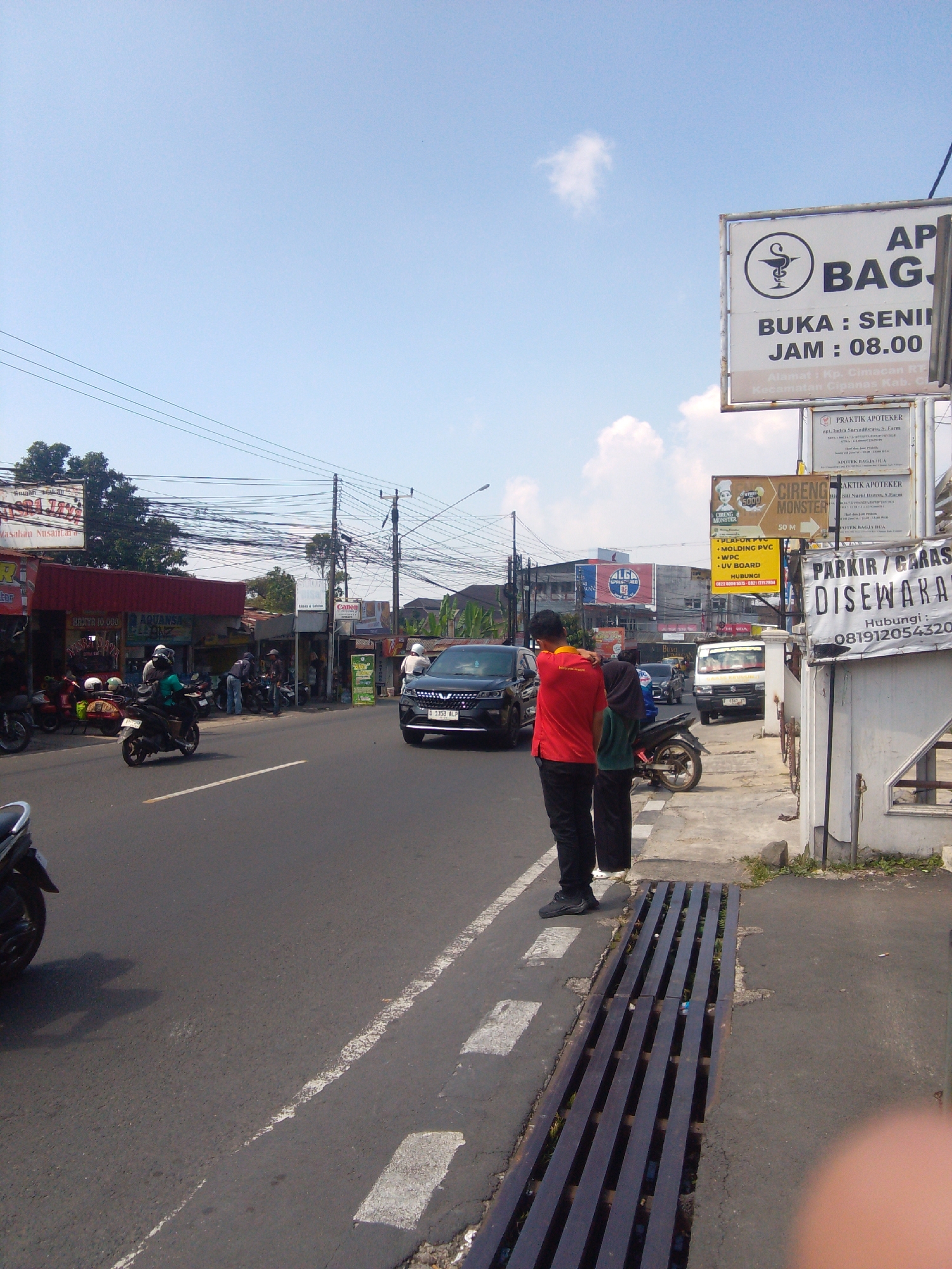 The image shows a street scene in Cianjur, Indonesia, on a bright, sunny day with a clear blue sky and some scattered clouds. A paved road runs through the center of the frame, with a white dashed line marking the lane. On the right side of the road, there is a sidewalk with a white wall and a metal grate. Several signs are visible, including one advertising "PARKIR / GARASI DISEWAKAN" (Parking / Garage for Rent) with a phone number. Further down the street, there are various shops and buildings, with some motorcycles parked along the sidewalk.
In the foreground, two individuals are standing on the sidewalk facing away from the camera. One person is wearing a red shirt and black pants, and the other is wearing a dark blue headscarf and a green shirt with black pants. A motorcycle is parked behind them.
On the road, several vehicles are present, including a black car, a white van, and various motorcycles. One motorcycle is being ridden by a person wearing a green shirt and a helmet. In the background, more buildings and power lines are visible, as well as trees. The overall impression is of a typical urban street in Indonesia.