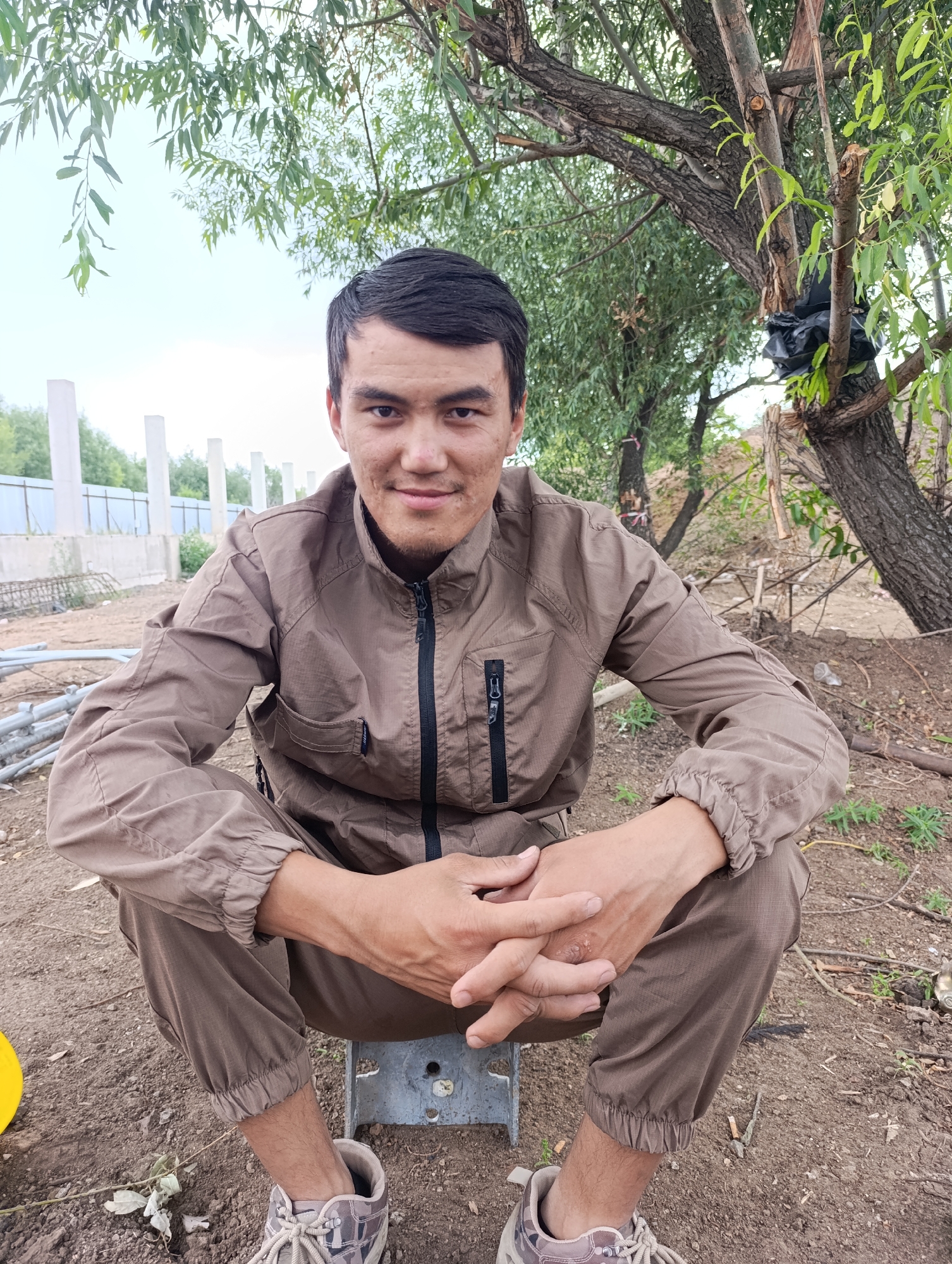 A young man is crouching on a metal stool outdoors, his hands clasped in his lap. He is wearing a brown zipped jacket and matching pants, with camouflage-patterned sneakers on his feet. He has dark hair and a light complexion, and he is looking directly at the camera with a slight smile. The background consists of dirt, scattered branches, and a tree with green leaves. In the distance, there are some white concrete pillars and a fence. The overall scene suggests an outdoor, possibly rural or industrial, setting.