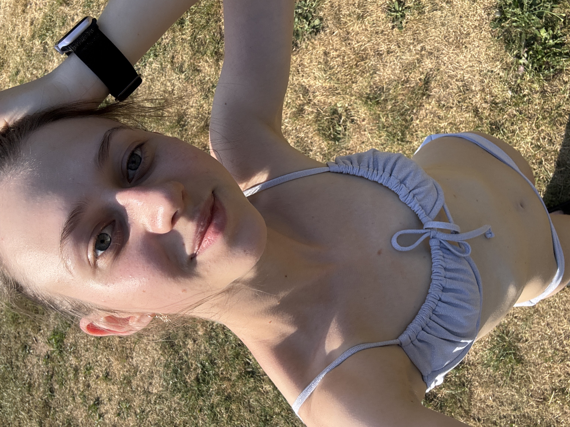 A young woman, with fair skin and light brown hair, is pictured outdoors, lying on dry grass. She is wearing a light grey, shimmery bikini. Her left arm is bent, resting her head, and a black smartwatch with a fabric band is visible on her wrist. Her right arm is extended upwards, with her hand near her head. The sunlight creates shadows on her skin, suggesting it is daytime with clear weather. The background consists of mostly dry, brown grass with some patches of green. The image is a selfie, taken from a high angle, with the woman's face close to the camera. Her expression is neutral, and she appears relaxed.