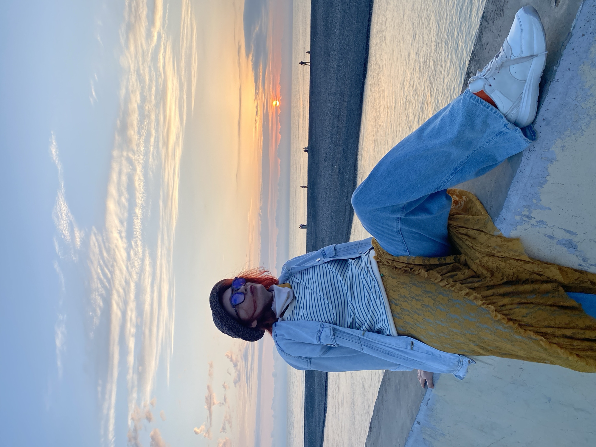 A woman is reclining on a concrete ledge with a view of the sunset over the sea in Sochi, Russia. She is wearing a black knit hat, blue sunglasses, a blue and white striped shirt, a light blue denim jacket, wide-legged blue jeans, white sneakers with orange socks showing, and a long yellow skirt. Her hair is red. The sun is setting over the water, visible in a gap in the clouds. There are a few figures standing in the distance on a dark beach or breakwater. The sky is light blue with some streaky clouds tinged with orange and yellow from the sunset.
