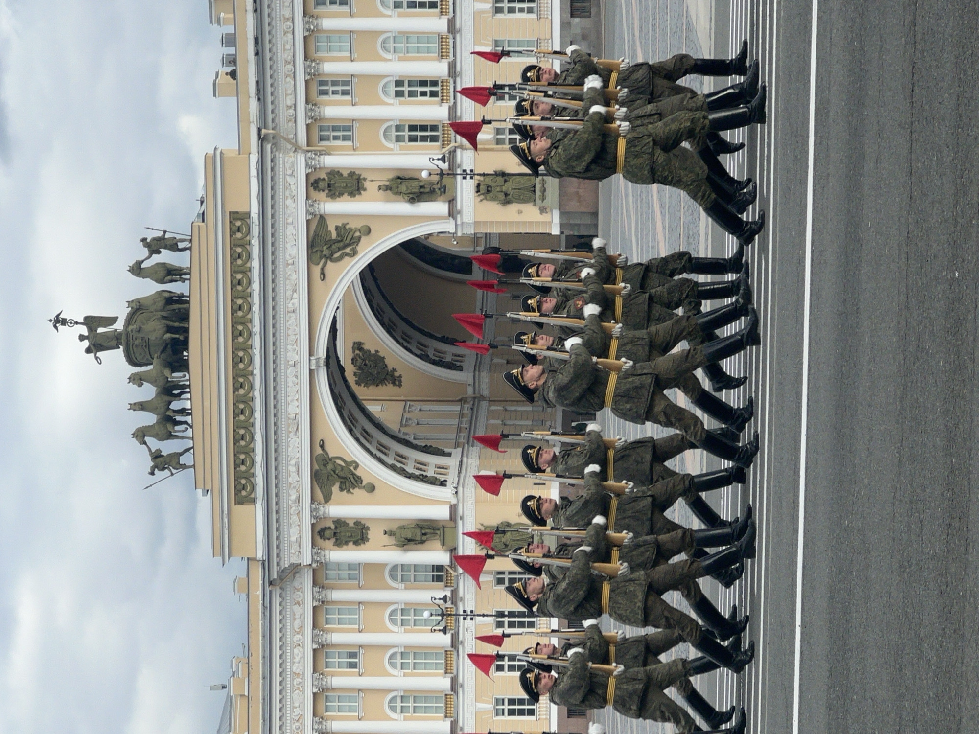 This image depicts a military parade or procession taking place on a paved road next to a grand architectural structure.
The primary subjects are a line of soldiers marching in formation from left to right across the frame. They are dressed in camouflage military uniforms, dark boots, and dark caps with insignia. Each soldier carries a rifle with a bayonet and a small, rectangular red flag attached near the muzzle. They appear to be marching in unison, their arms bent at the elbow, holding their rifles in a formal parade stance.
To the left of the marching soldiers stands a large, ornate, pale yellow neoclassical building with numerous white columns and arched windows. The building features an impressive archway through which more of the structure is visible. Adorning the roofline and façade are several statues and relief sculptures. Most notably, a large quadriga (a chariot drawn by four horses) with a figure driving it is prominently positioned on the roof of the building, accompanied by other standing figures. The sky above is cloudy, suggesting an overcast day. The road where the soldiers march is dark asphalt with white lines painted on it. No discernible text is visible in the image.