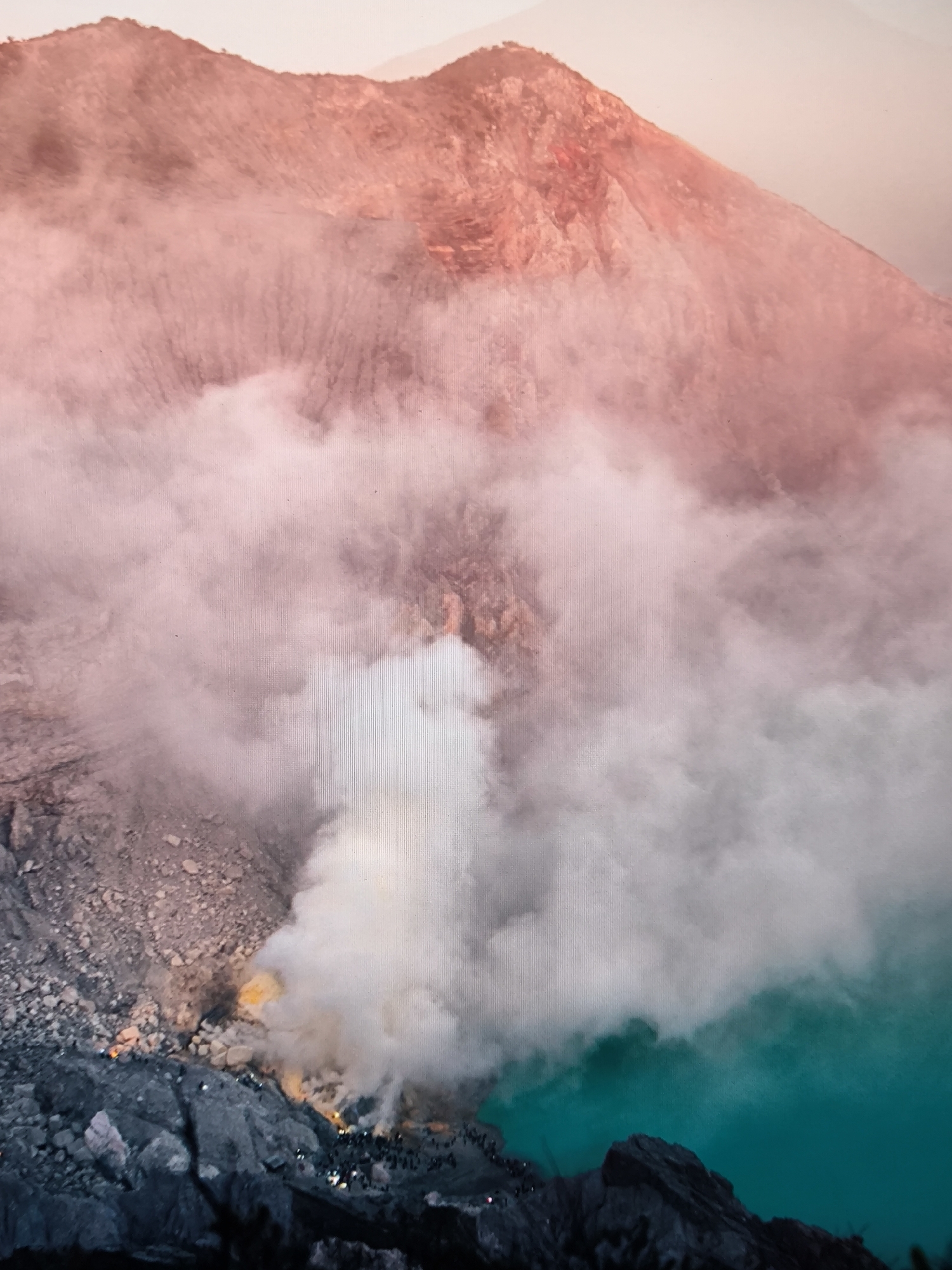 The scene depicts an active volcanic crater. In the upper portion, distant, reddish-pink mountainous terrain forms the crater rim, partially obscured by diffuse, pale pinkish-white atmospheric haze or smoke. The central and lower-left areas are dominated by a massive, billowing plume of white and light grey smoke or gas, rising from the crater floor.
At the base of this smoke plume, on rugged, dark grey and brown rocky terrain in the lower-left quadrant, numerous small human figures are discernible. These individuals, estimated to be several dozen to over a hundred, are clustered along the rocky slopes and edges. Many appear to be equipped with artificial light sources, such as headlamps, visible as small, bright white or yellow dots against the darker ground. Adjacent to these figures, distinct bright yellow and orange glows indicate areas of burning sulfur or intense sulfur deposits.
In the lower-right quadrant, a large body of intensely turquoise-colored water, consistent with an acidic crater lake, occupies a significant portion of the view. Dark, jagged rock formations define the immediate foreground at the water's edge. The overall lighting suggests either dawn, dusk, or a nighttime scene illuminated by ambient light and artificial sources within the crater. No specific city is identifiable; the location is an active volcanic crater environment.