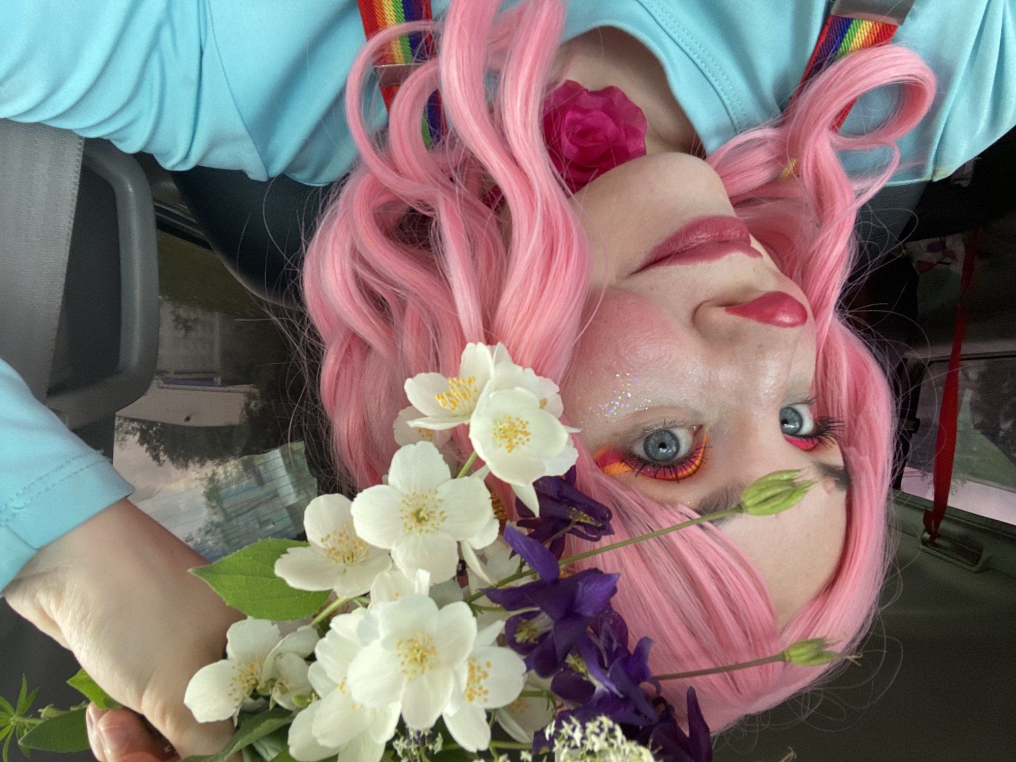 A close-up, profile shot shows a person with bright pink, wavy hair and clown makeup. The makeup includes glitter on the cheeks, bright orange and pink eyeshadow, a red clown nose, and pink lipstick. They are wearing a light blue t-shirt with rainbow-colored suspenders. A crown of white jasmine flowers and purple delphiniums is woven into their hair. The person is inside a vehicle, as indicated by the car interior visible in the background. The scene does not provide any specific information to locate it in Chaikovsky, Russia.