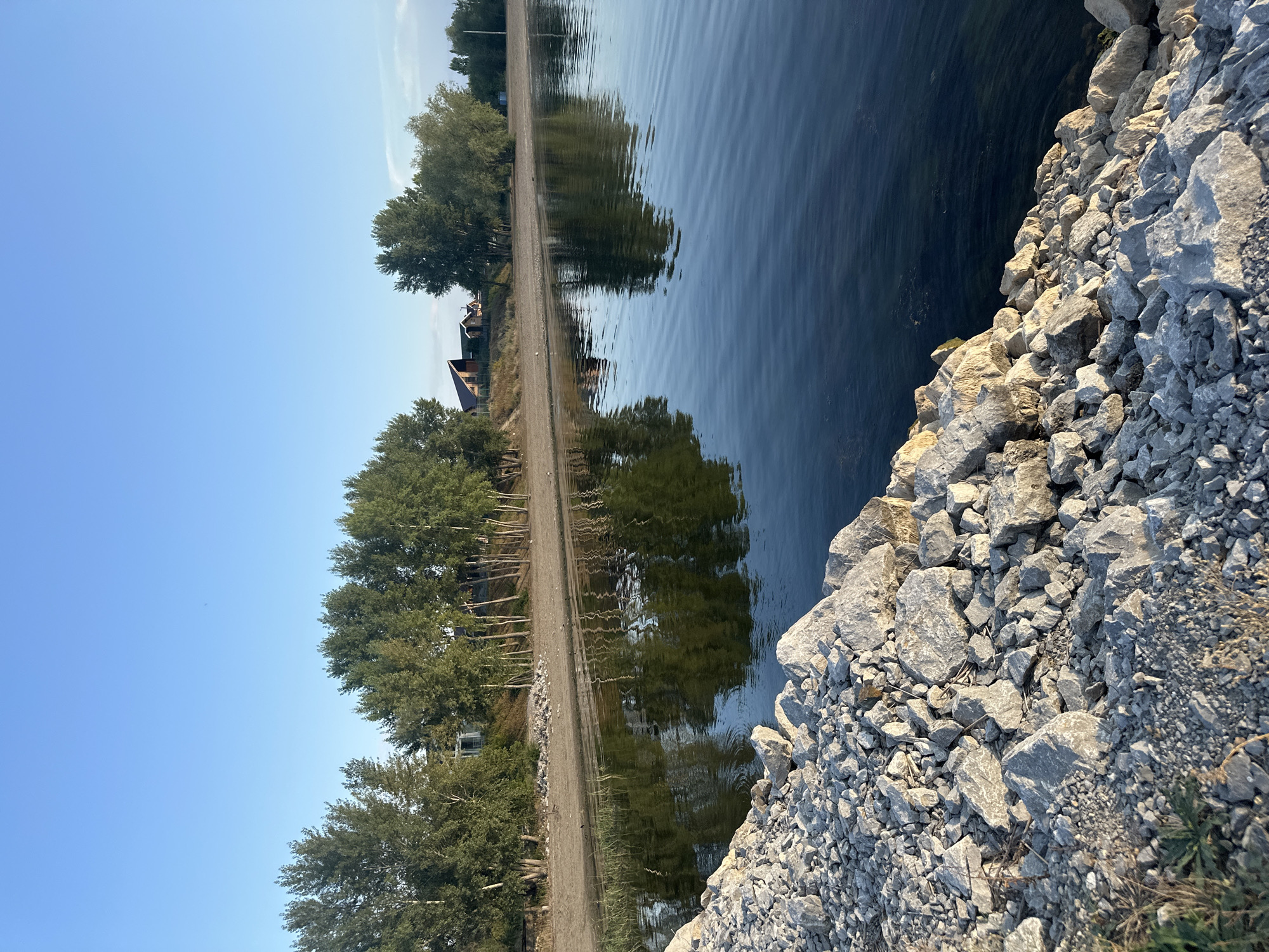 A serene, wide-angle shot captures a body of water with a tree-lined shore and a rocky embankment in the foreground. The water, a deep blue, reflects the clear sky and the lush green trees with a subtle ripple effect. On the sandy shore, a line of trees stands tall, their leaves a vibrant green. Behind them, several houses with angled roofs are visible, suggesting a residential area. The foreground is dominated by a rough embankment constructed of various sizes of gray and white rocks. The scene is bathed in the warm light of what appears to be late afternoon or early morning, casting gentle shadows. The overall impression is one of peaceful natural beauty, with hints of human habitation on the horizon. The location is identified as Energetik, Russia.