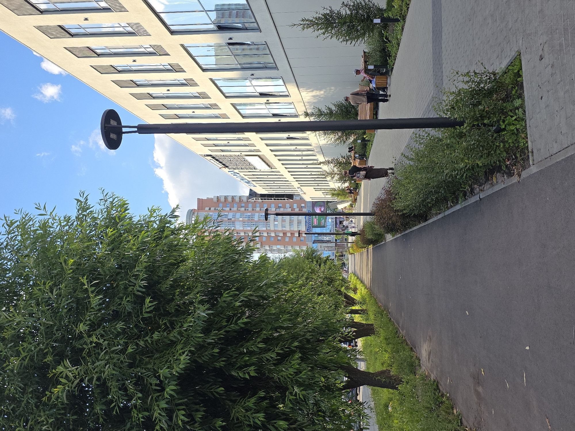 The image depicts a modern urban scene with a pedestrian walkway alongside a tree-lined street and a contemporary building. Tall, leafy trees with dense green foliage are positioned on the left side of the image, casting shadows on the asphalt path. Beyond the trees, a street with moving cars is visible. On the right side of the walkway is a long, beige building with a facade featuring numerous large windows and a clean, geometric design. The walkway itself is paved with light gray interlocking bricks. Several people are present on the walkway and sitting on benches. Two individuals, one in a brown shirt and one in a plaid shirt, are seated on a wooden bench. Another person in a long dark dress walks away from the camera, while a man in a light brown shirt and dark pants walks toward the building. A lamppost with a modern circular head stands on the walkway. In the background, a tall residential building with visible windows and balconies can be seen, along with smaller evergreen trees planted along the building's exterior. The sky is blue with scattered white clouds. Based on architectural style and street landscaping, this scene is characteristic of modern development in cities like Chelyabinsk, Russia.
