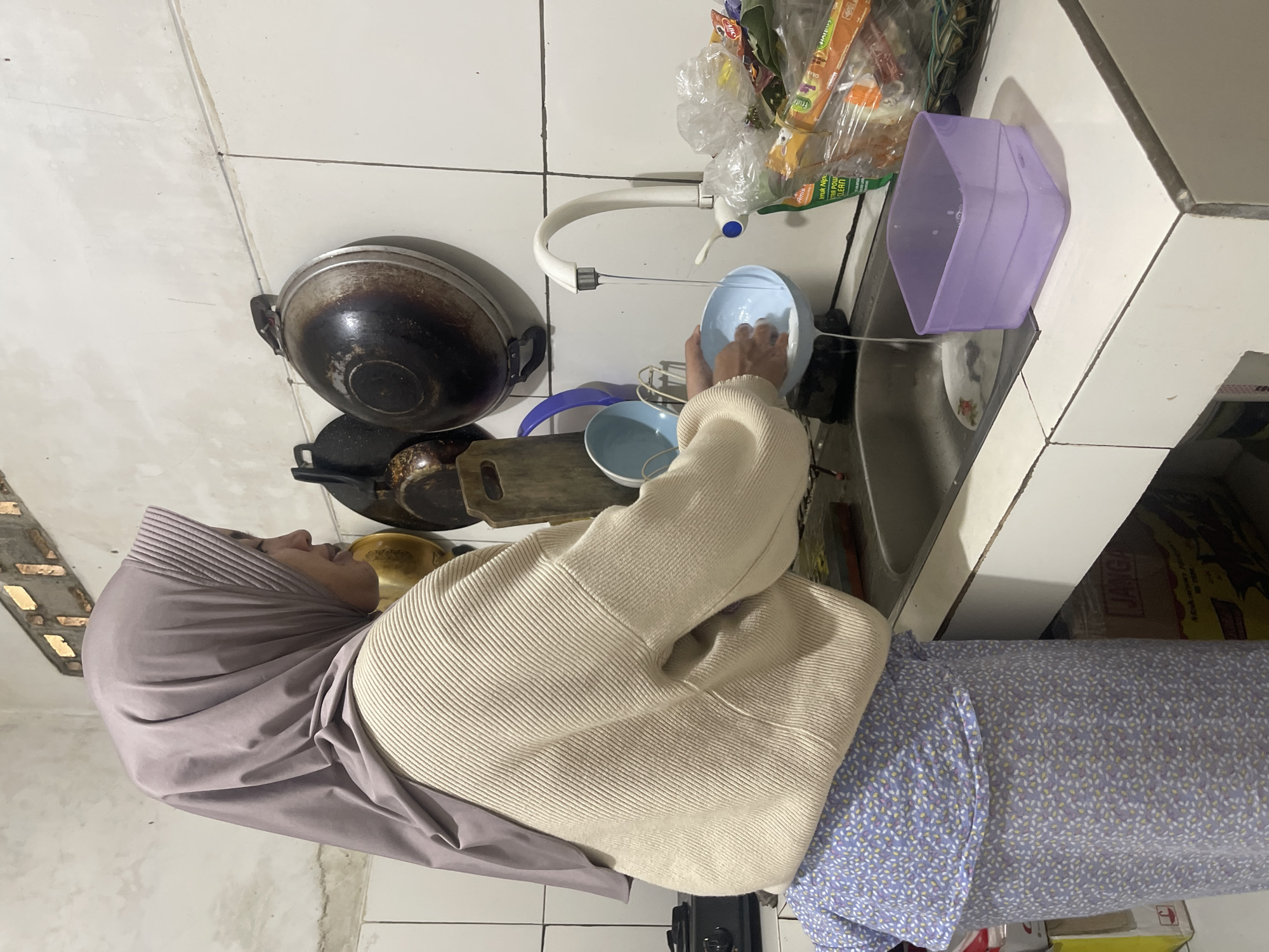 A woman wearing a light purple hijab and a cream-colored ribbed sweater is washing a light blue bowl in a stainless steel sink. Her patterned purple skirt is visible from the waist down. The sink is part of a white countertop in what appears to be a kitchen. Behind the sink, a white faucet is running water into the bowl. Hanging on the tiled wall are several dark-colored pots. To the right of the sink, plastic-wrapped items, possibly food or cleaning supplies, are stacked. A light purple plastic container sits on the countertop next to the sink. The scene is indoors, likely in Gunung Sindur, Indonesia, based on the context provided.