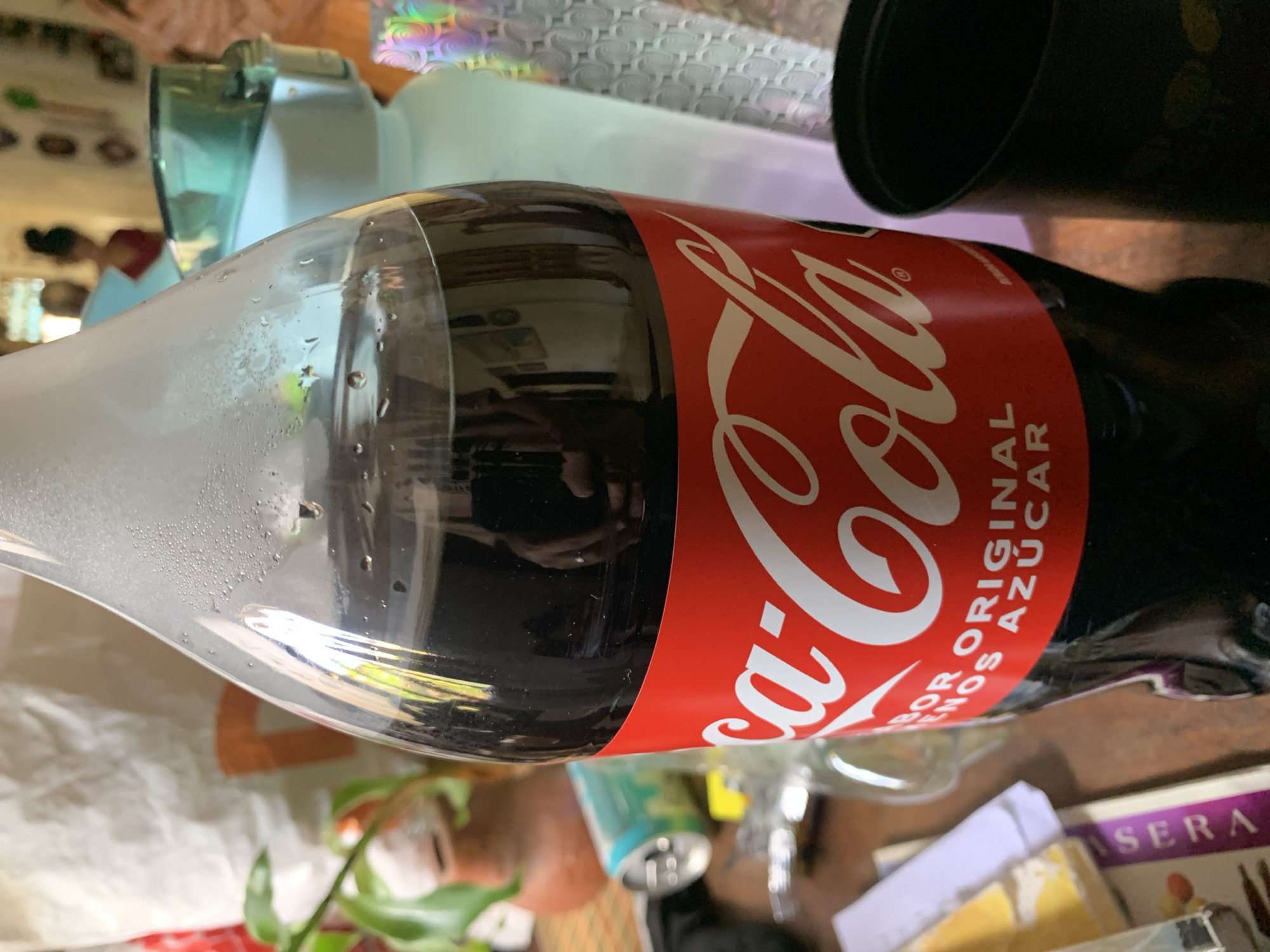 This media file, captured in Santa Ana, Venezuela, presents a close-up view of a cold, plastic Coca-Cola bottle. The bottle is lying horizontally on what appears to be a wooden surface, showing significant condensation, indicating it has recently been removed from refrigeration.
The central subject is the Coca-Cola bottle, prominently featuring its iconic red label with the white "Coca-Cola" script logo. Visible Spanish text on the label includes "SABOR ORIGINAL" (Original Flavor) and below it, "AZÚCAR" (Sugar), identifying it as the regular, sugared version of the beverage.
Reflections on the shiny surface of the bottle reveal parts of the immediate environment, including what looks like a hand holding a smartphone, presumably the photographer. The overall scene suggests an indoor setting, likely a kitchen or dining area.
In the background, various out-of-focus objects are visible: a light blue or green plastic jug or container, a black cup or container, and a material with a holographic or patterned texture. Further down, there's a brown potted plant, a crumpled white object (possibly a bag or paper), and a glimpse of another beverage can. On the lower right, part of a package with the text "SERA" is partially seen. The lighting is bright, suggesting daytime. The image captures a casual moment with the cold drink in its immediate surroundings.
